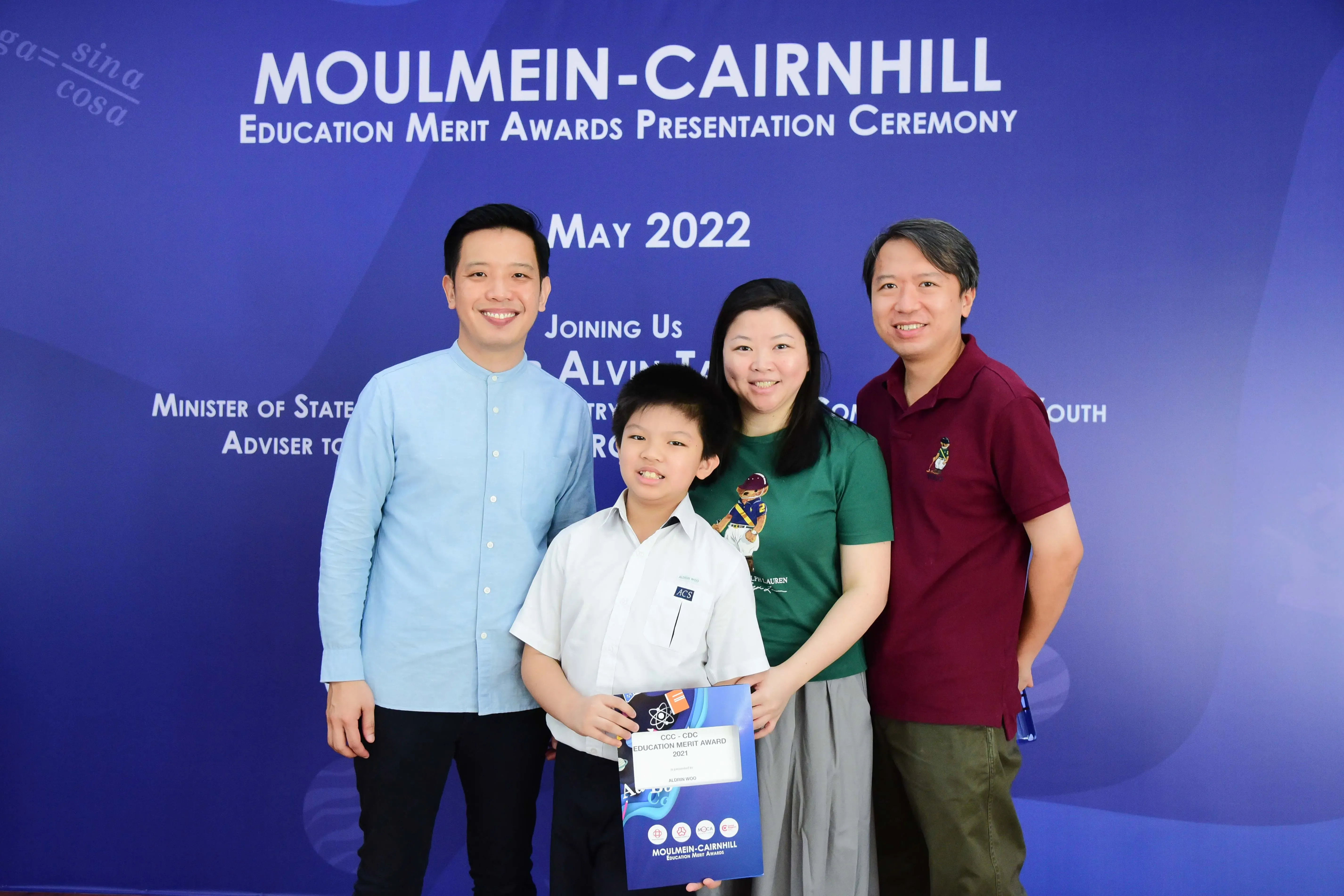 A boy holding an Education Merit Award certificate poses with three adults at the Moulmein-Cairnhill ceremony, May 2022.