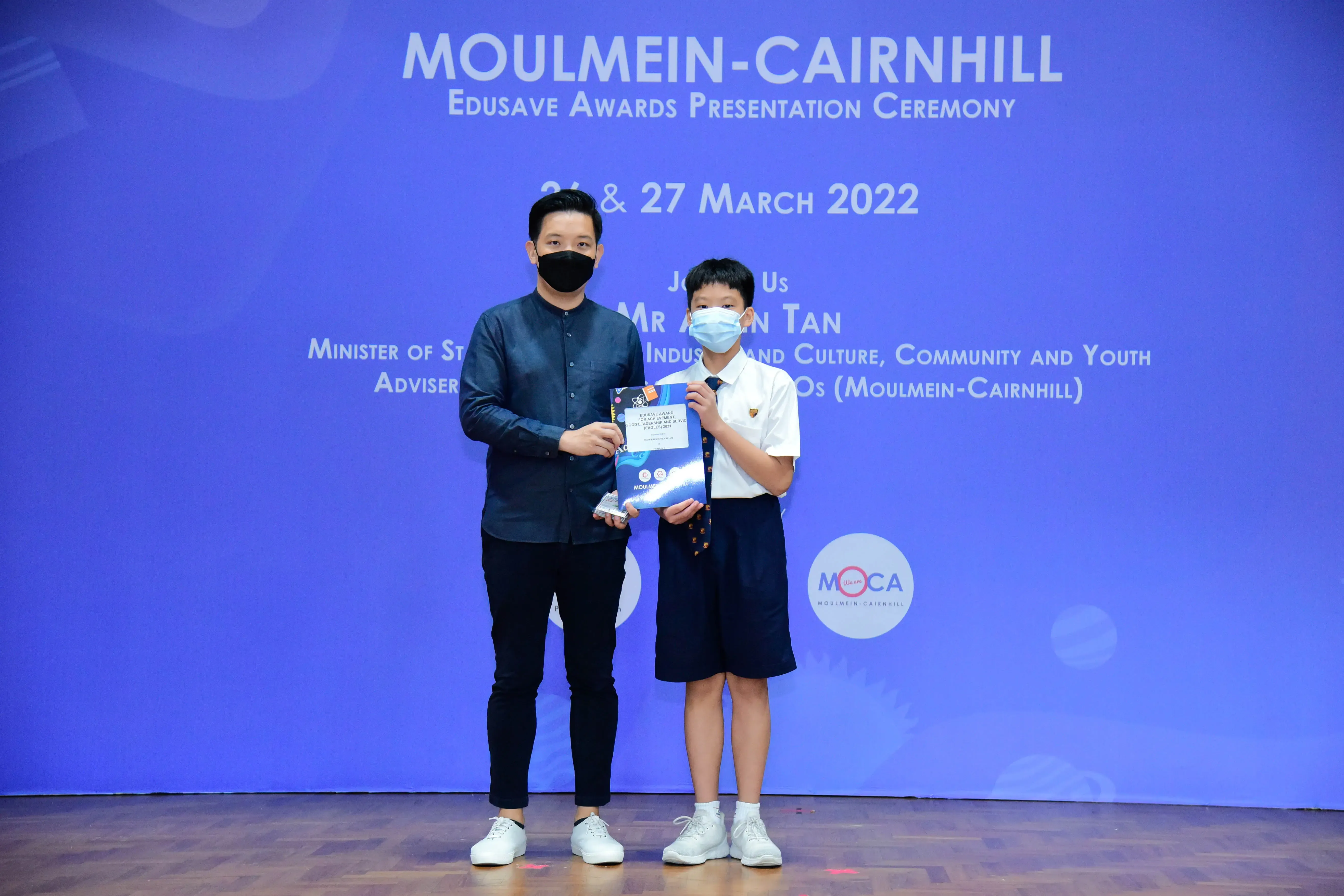 An adult and a young student in school uniform hold a certificate at the Moulmein-Cairnhill Edusave Awards Ceremony 2022.