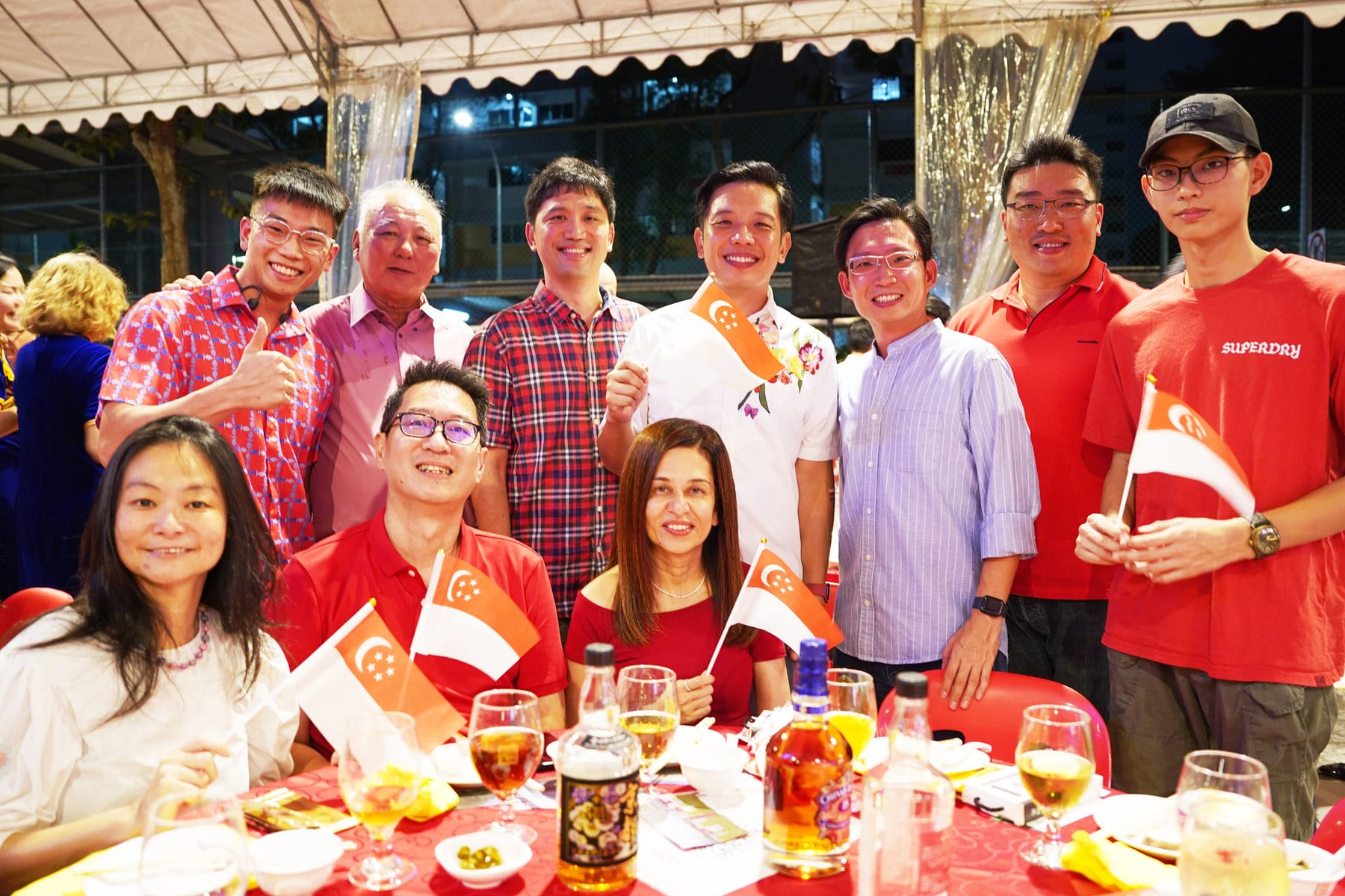 Group of smiling people at an outdoor evening event holding small Singapore flags with drinks on a red-clothed table.