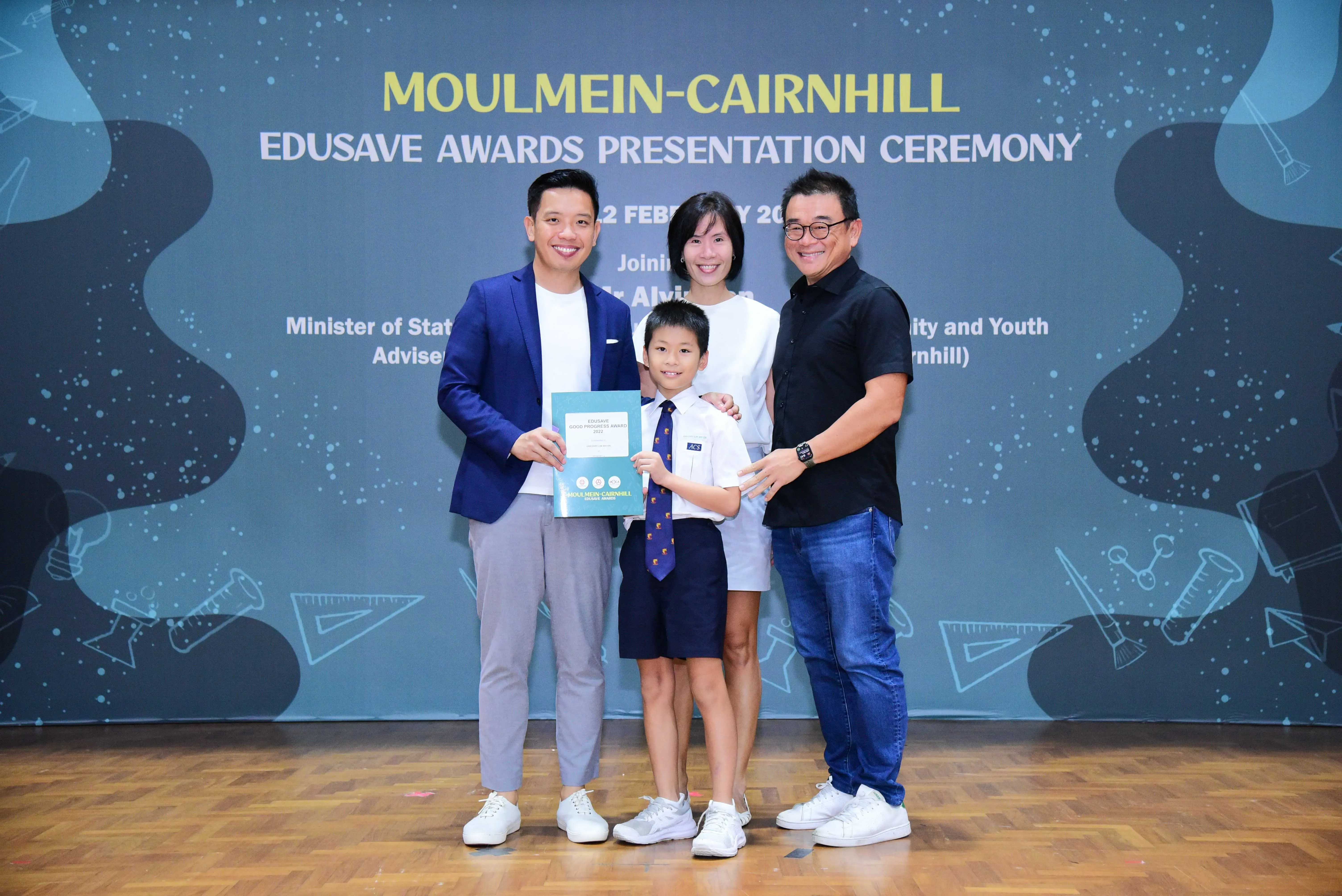 Four people pose at the Moulmein-Cairnhill Edusave Awards Presentation Ceremony, including a boy in school uniform holding a certificate.
