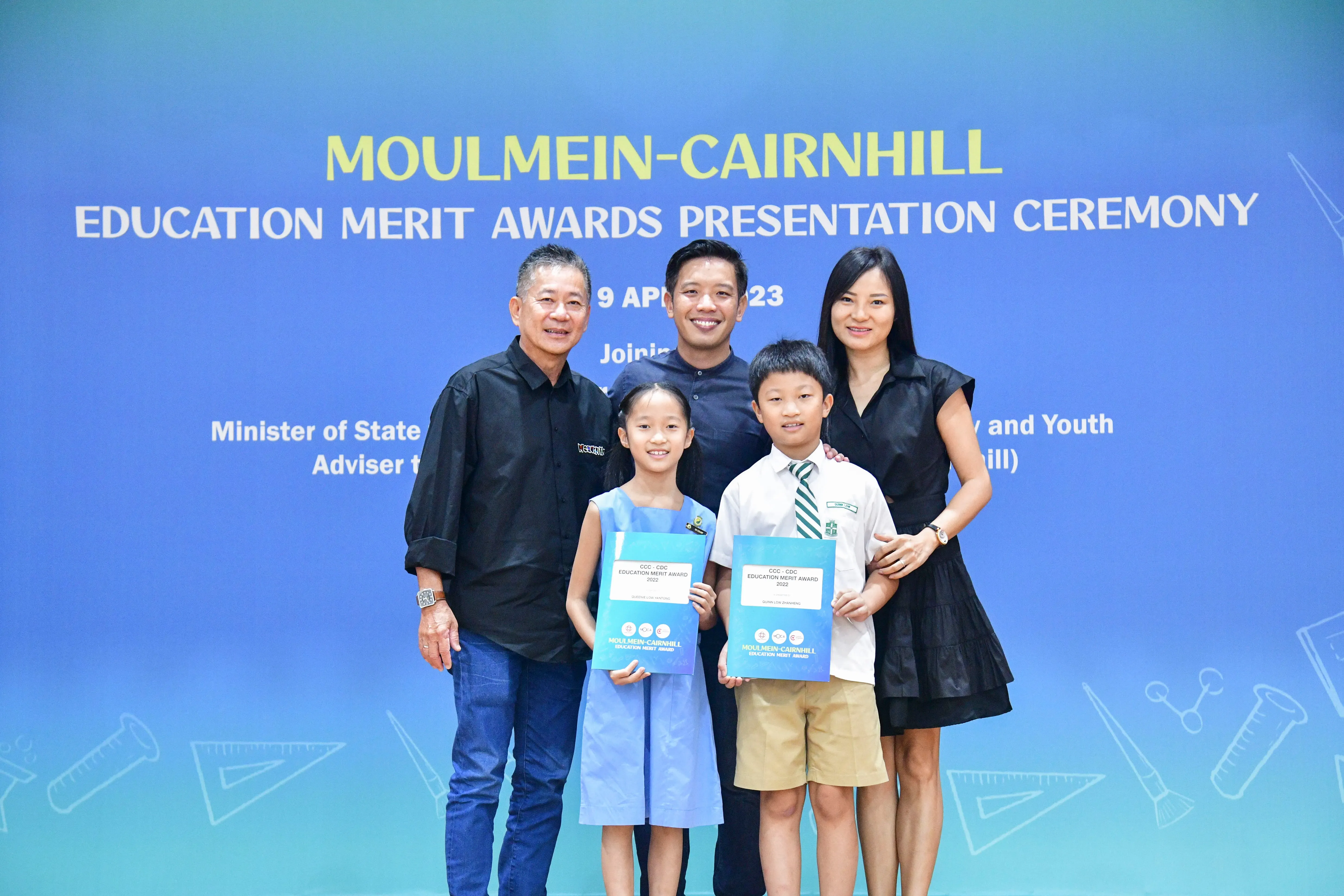 Five people pose before a Moulmein-Cairnhill Education Merit Awards banner; two children hold award certificates.