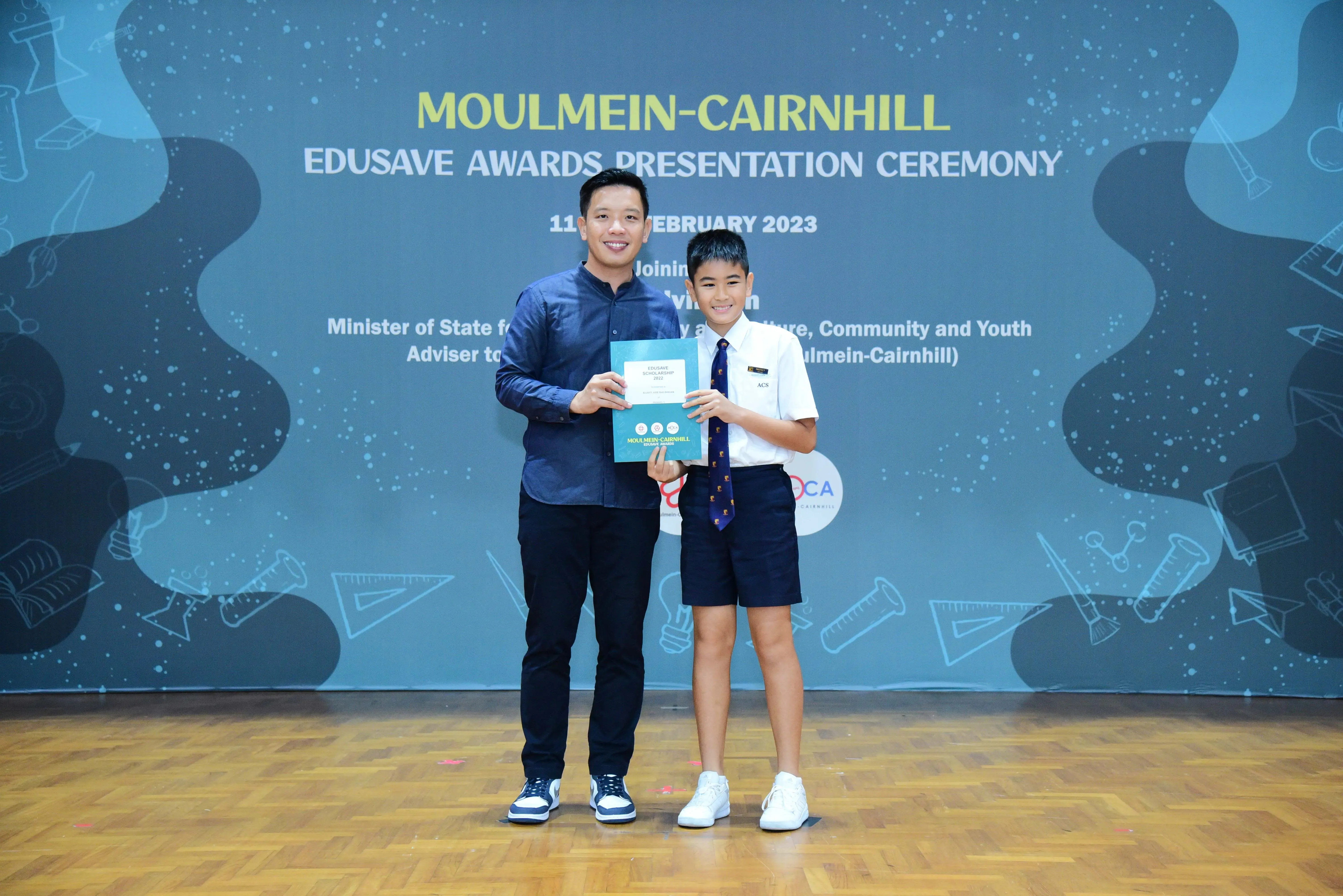 A man and a young student in school uniform hold a certificate at the Moulmein-Cairnhill Edusave Awards Ceremony 2023.