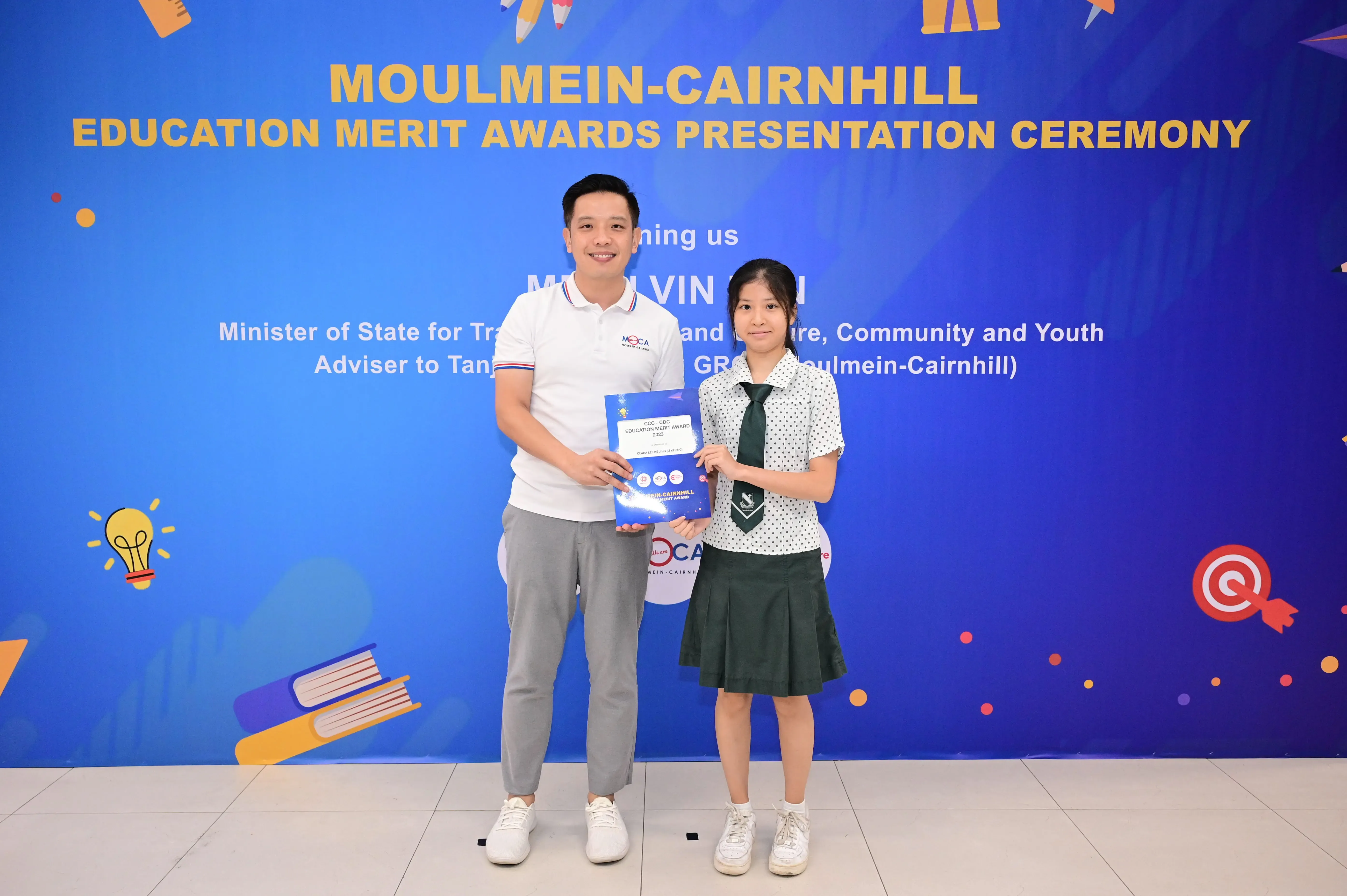 A man and a young girl in school uniform hold a blue award certificate at the Moulmein-Cairnhill Education Merit Awards.