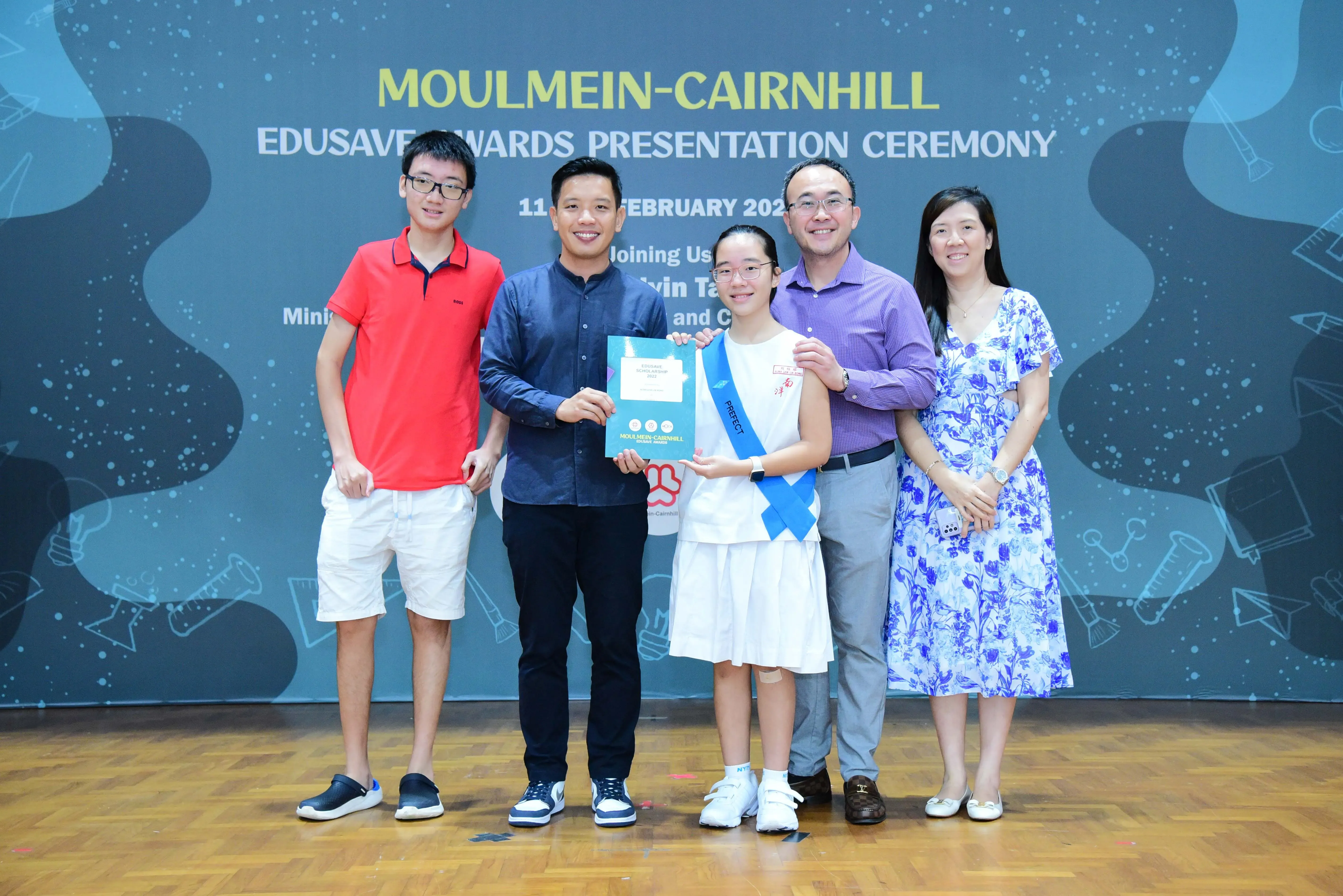 Five people pose at the Moulmein-Cairnhill Edusave Awards Presentation Ceremony, with a girl holding an award certificate.