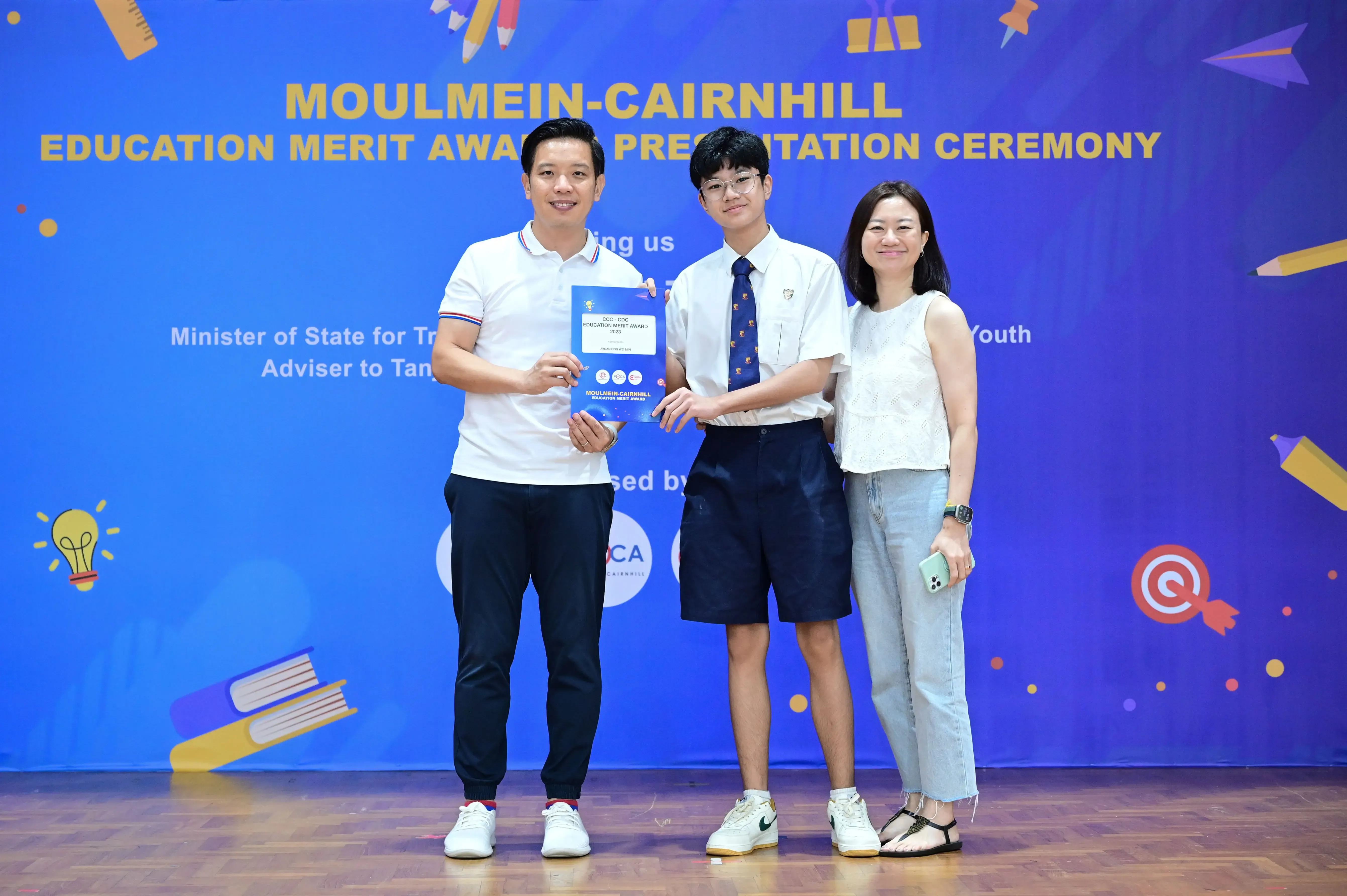 Three people stand together holding a certificate at the Moulmein-Cairnhill Education Merit Award Presentation Ceremony.