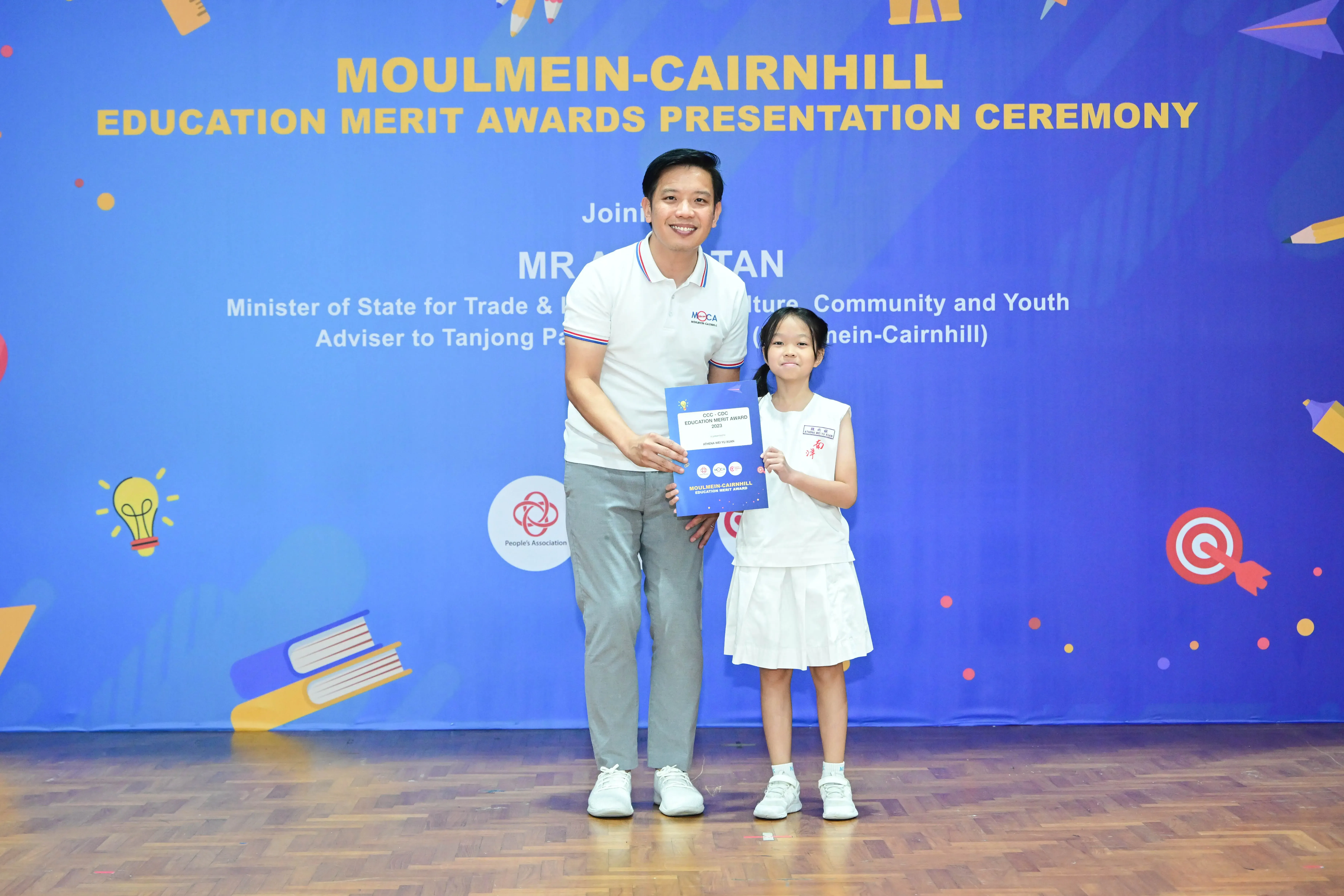 A man and a young girl hold a certificate in front of a Moulmein-Cairnhill Education Merit Awards banner.