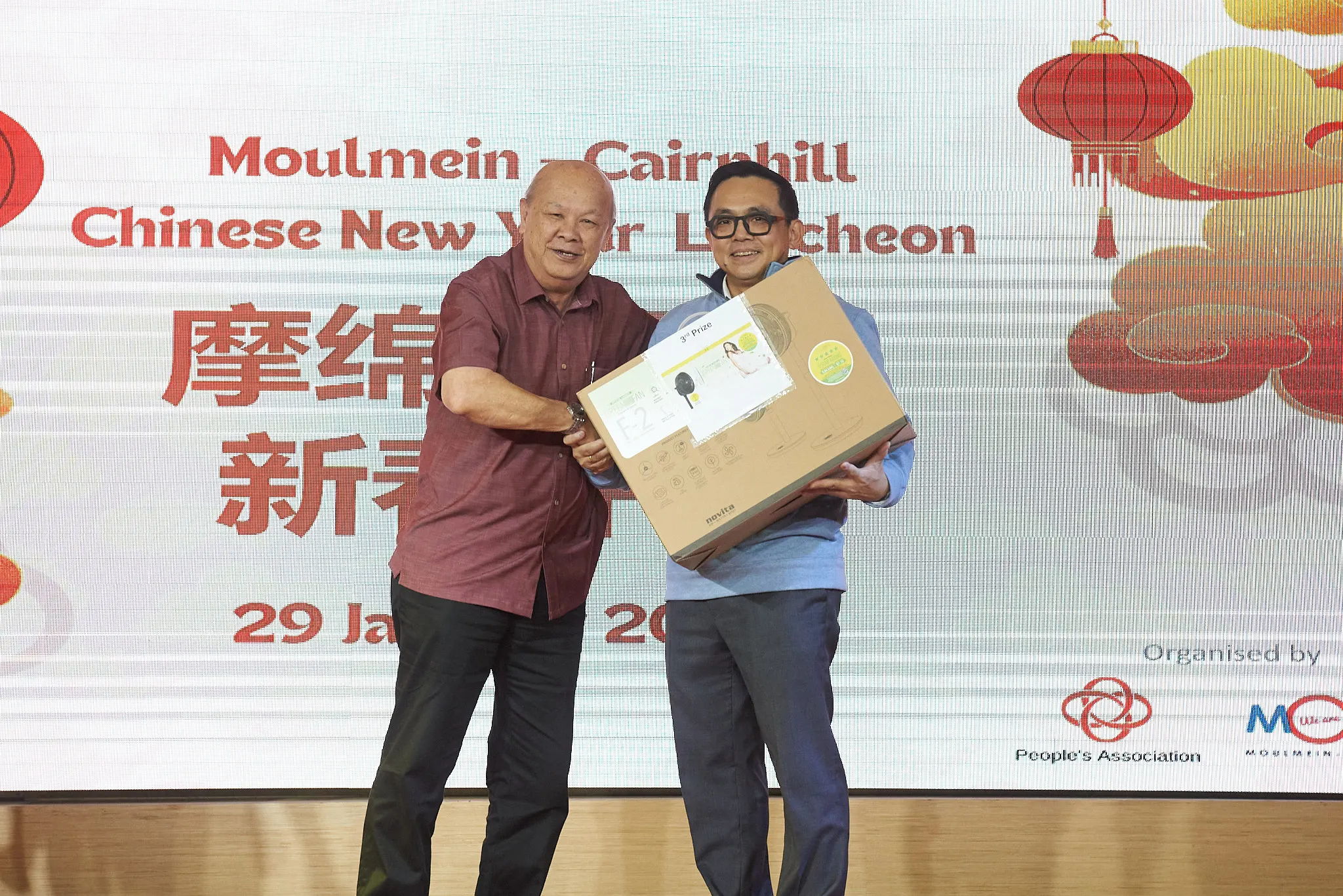 Two men hold a boxed prize on stage at the Moulmein-Cairnhill Chinese New Year Luncheon event.
