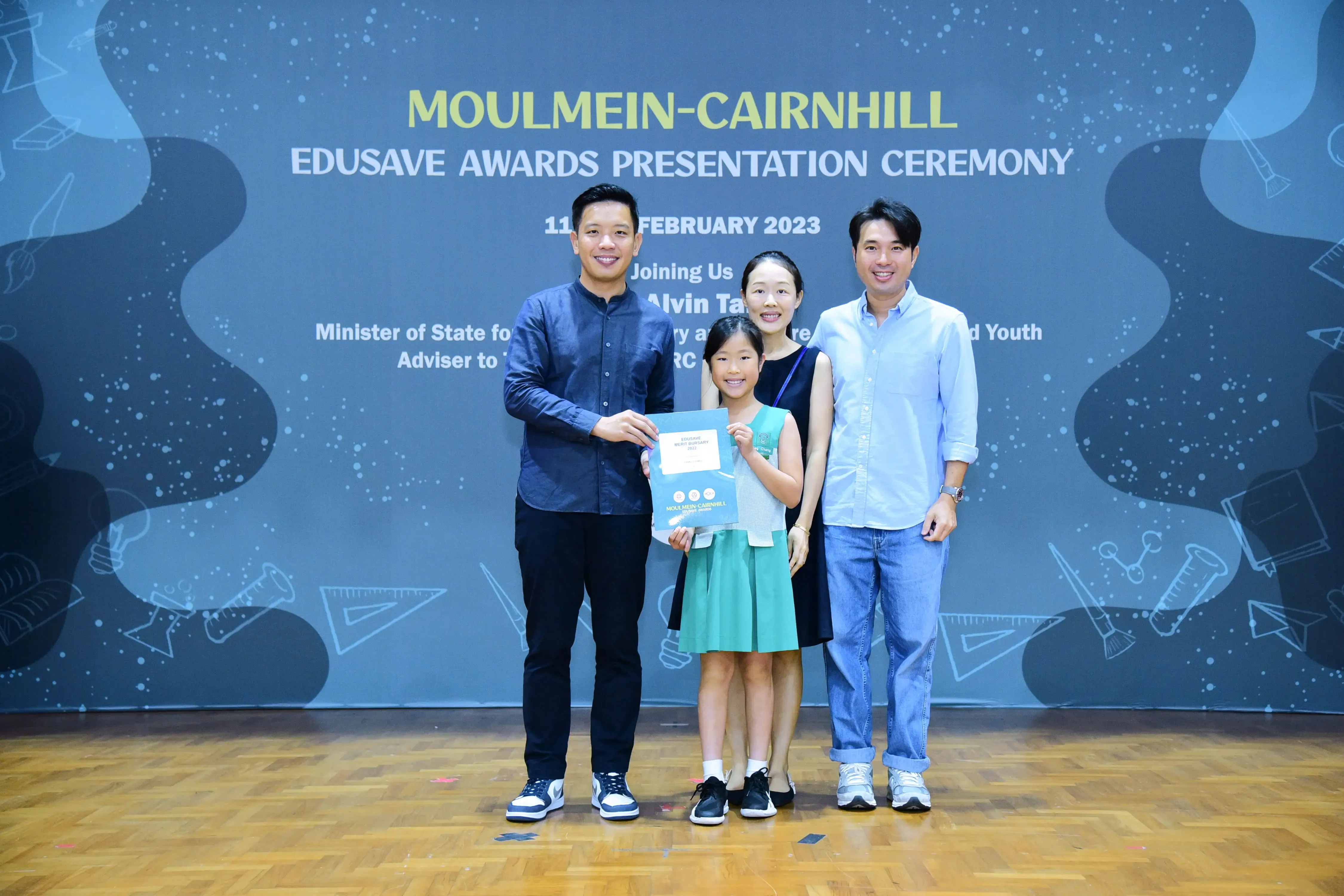 Four people pose at the Moulmein-Cairnhill Edusave Awards Presentation Ceremony, with a girl holding a certificate.