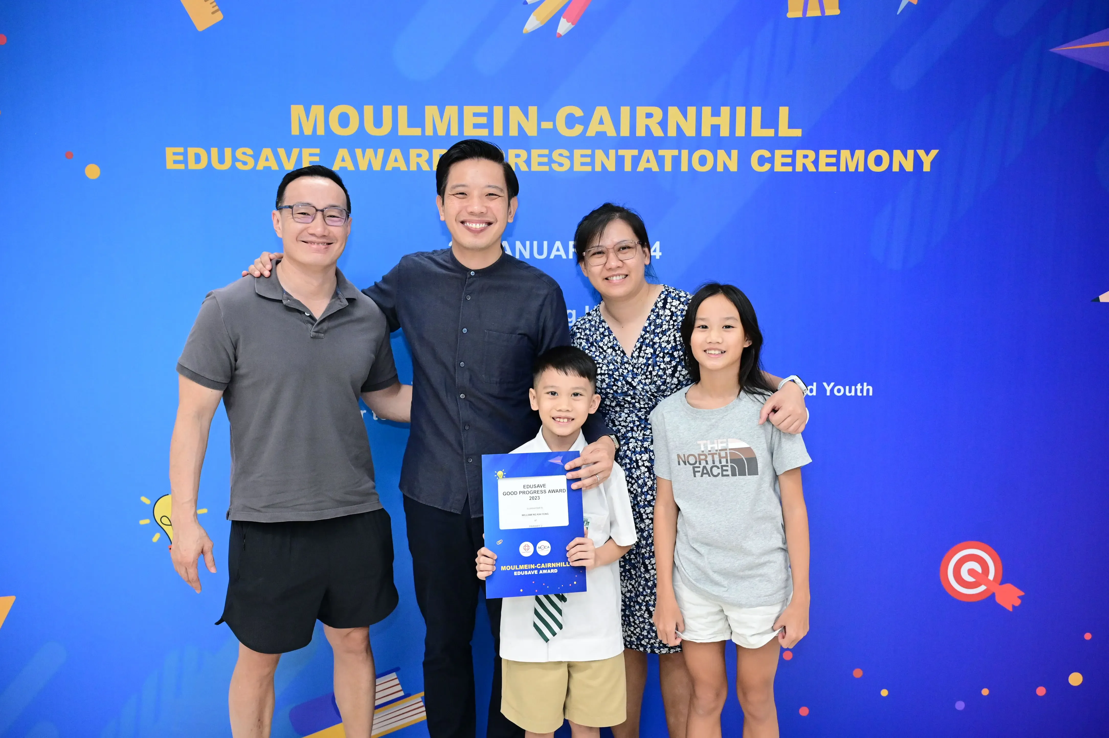 Five people smile in front of a blue Moulmein-Cairnhill Edusave Award Presentation Ceremony banner, boy holds certificate.