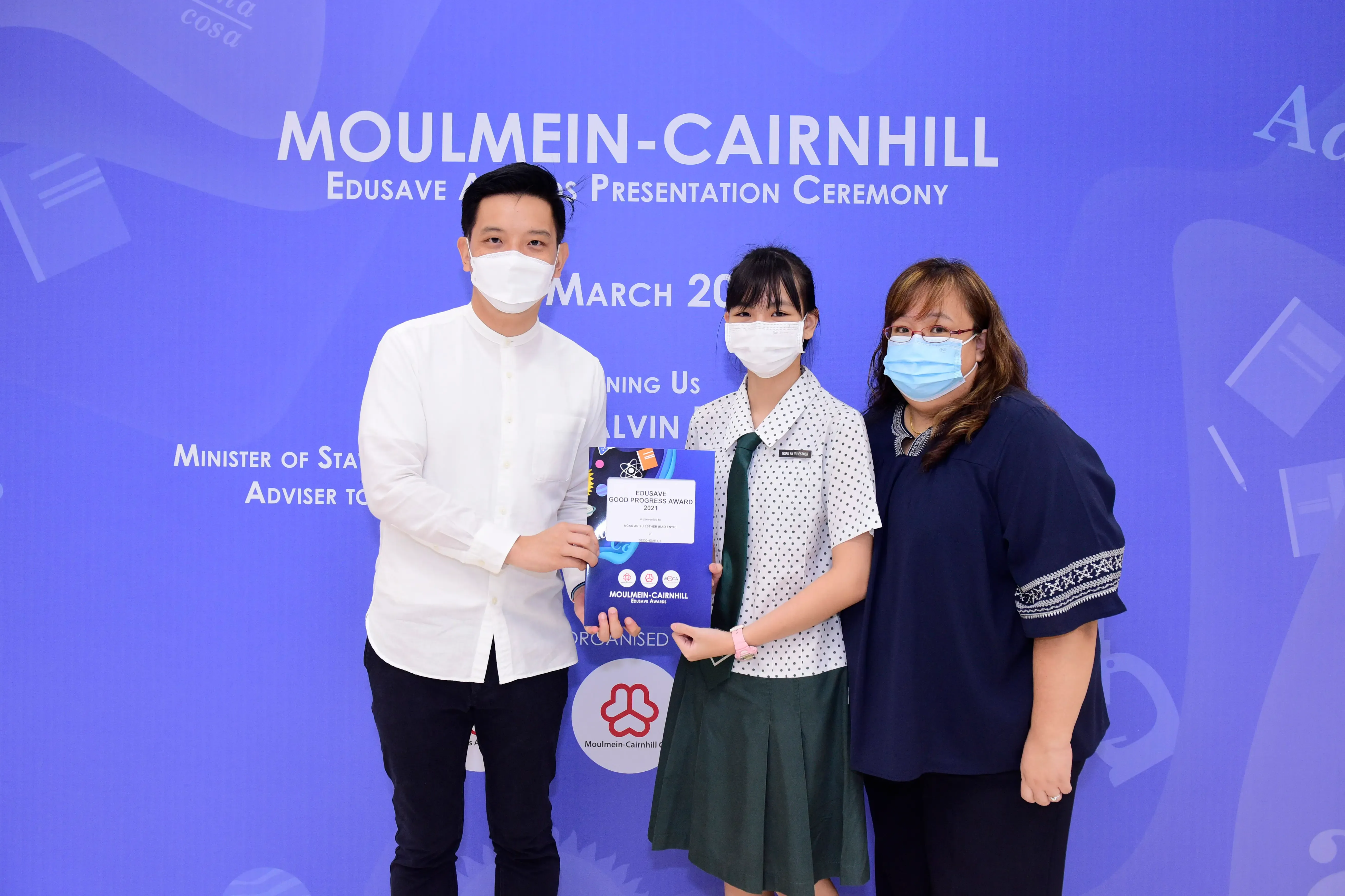 Three masked people pose at the Moulmein-Cairnhill Edusave Awards Ceremony, with a student holding an award certificate.