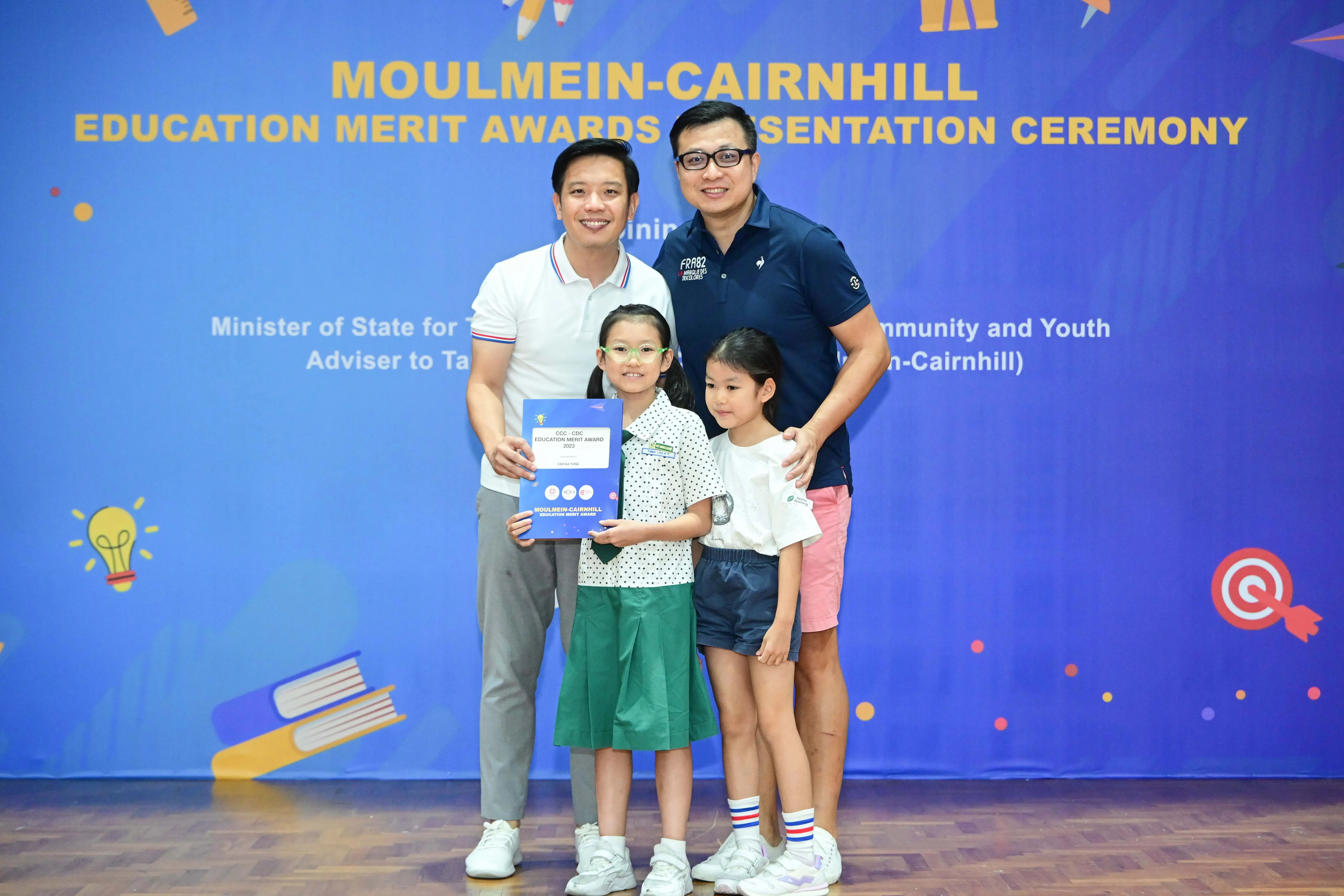 Two men and two young girls pose at the Moulmein-Cairnhill Education Merit Awards Presentation Ceremony, one girl holding a certificate.