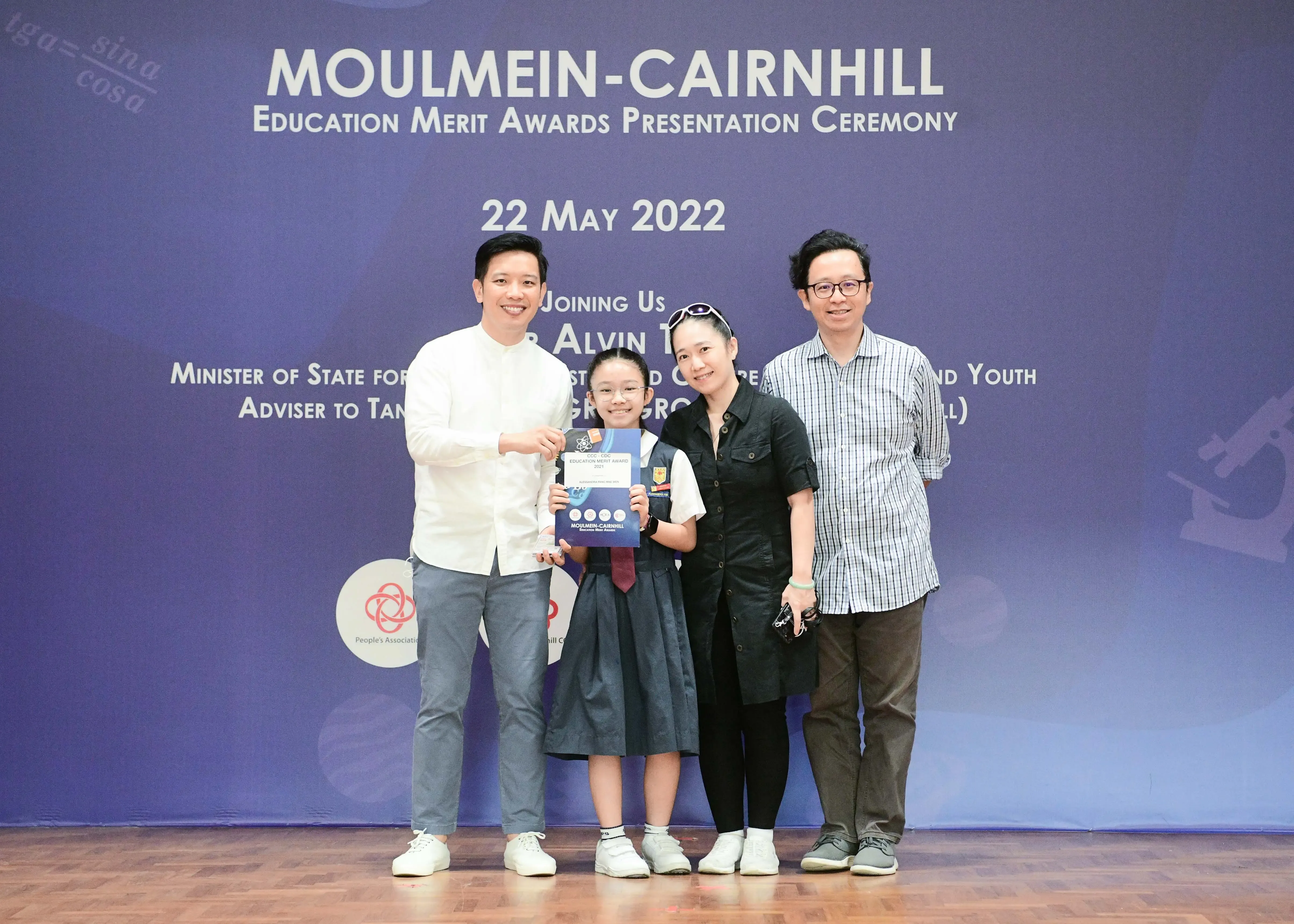 Four people pose at the Moulmein-Cairnhill Education Merit Awards ceremony on 22 May 2022, with a girl holding a certificate.