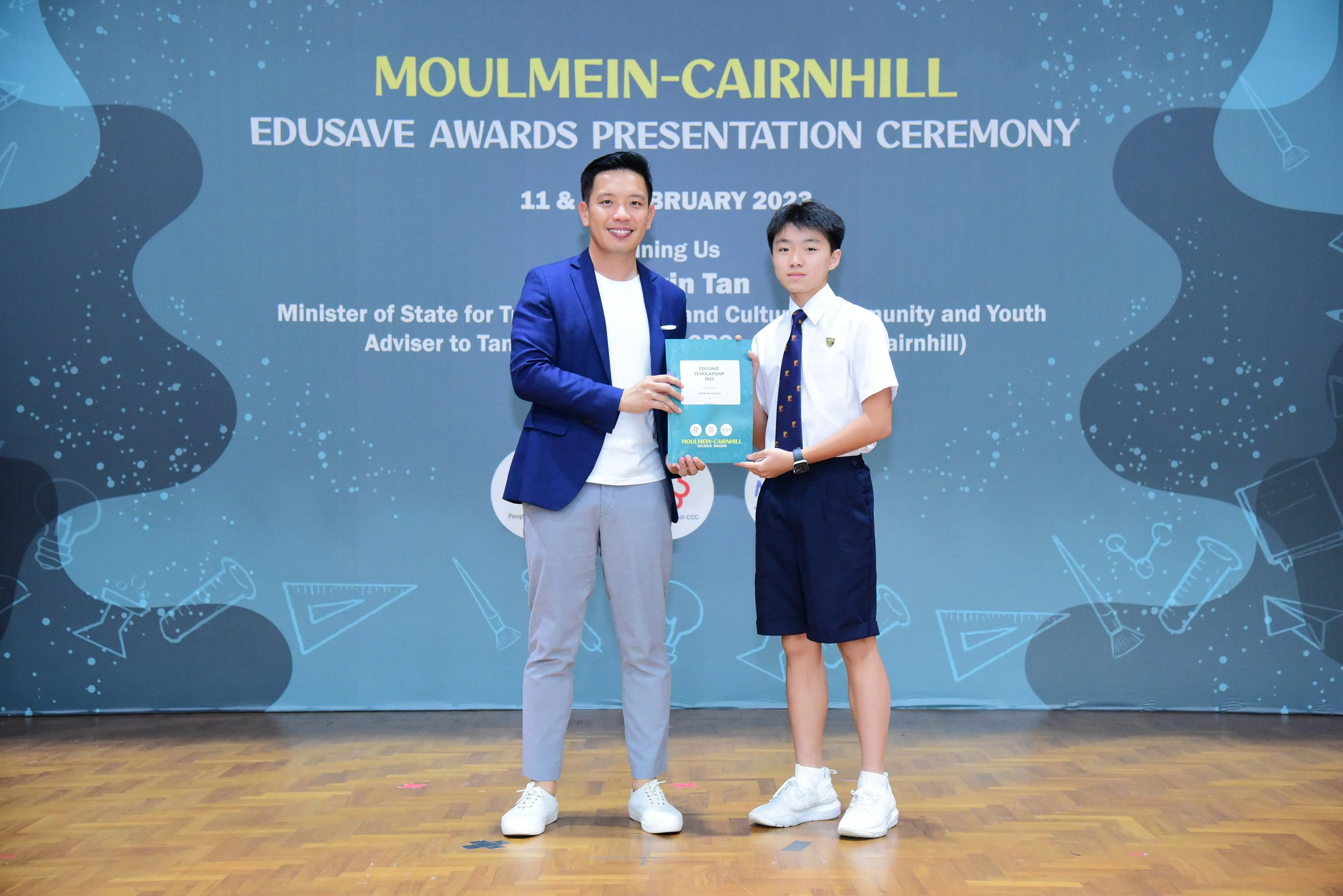 An adult in a blue blazer presents an award to a uniformed student at the Moulmein-Cairnhill Edusave Awards Ceremony 2023.