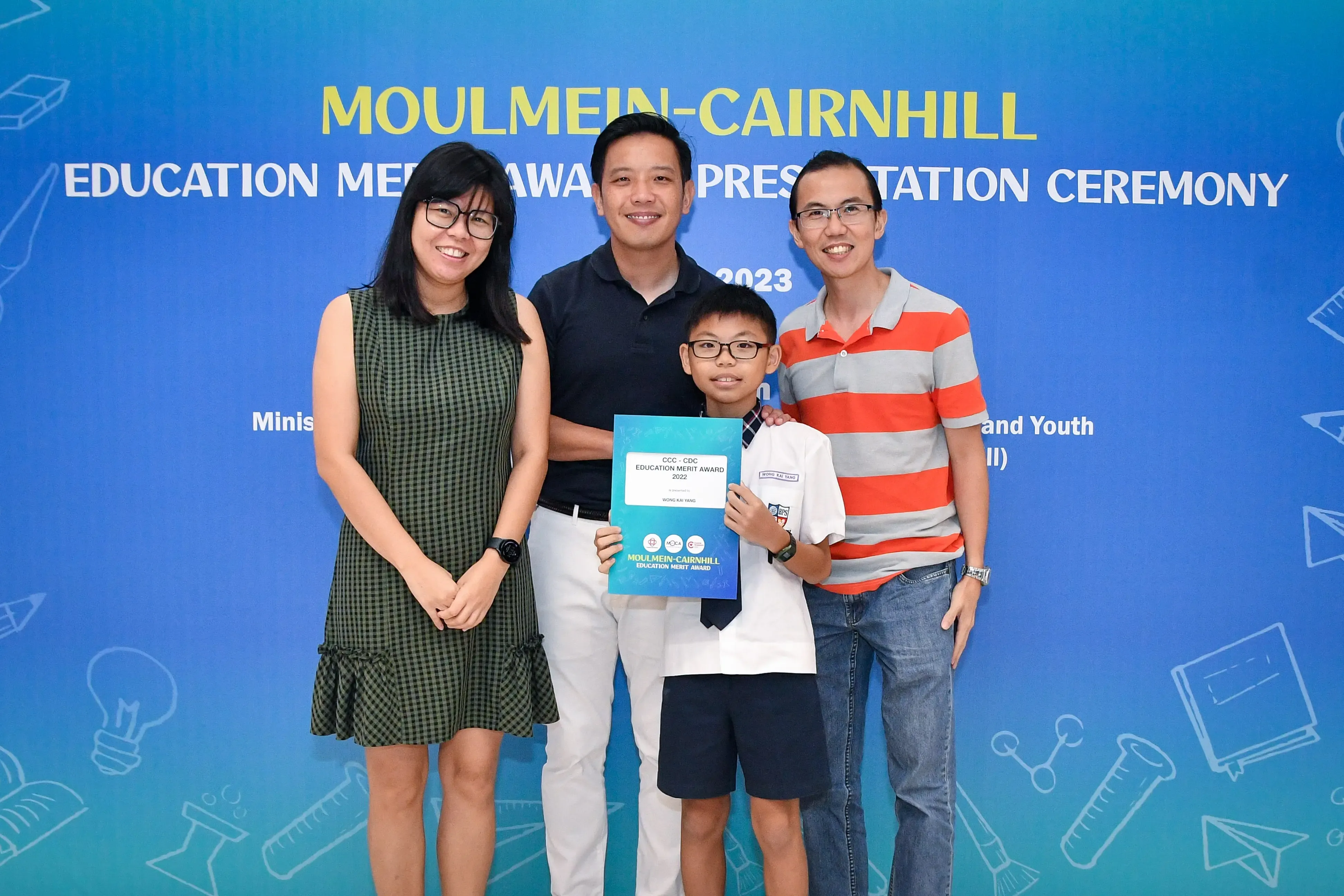 A boy holding an education merit award certificate poses with three smiling adults at the 2023 Moulmein-Cairnhill ceremony.