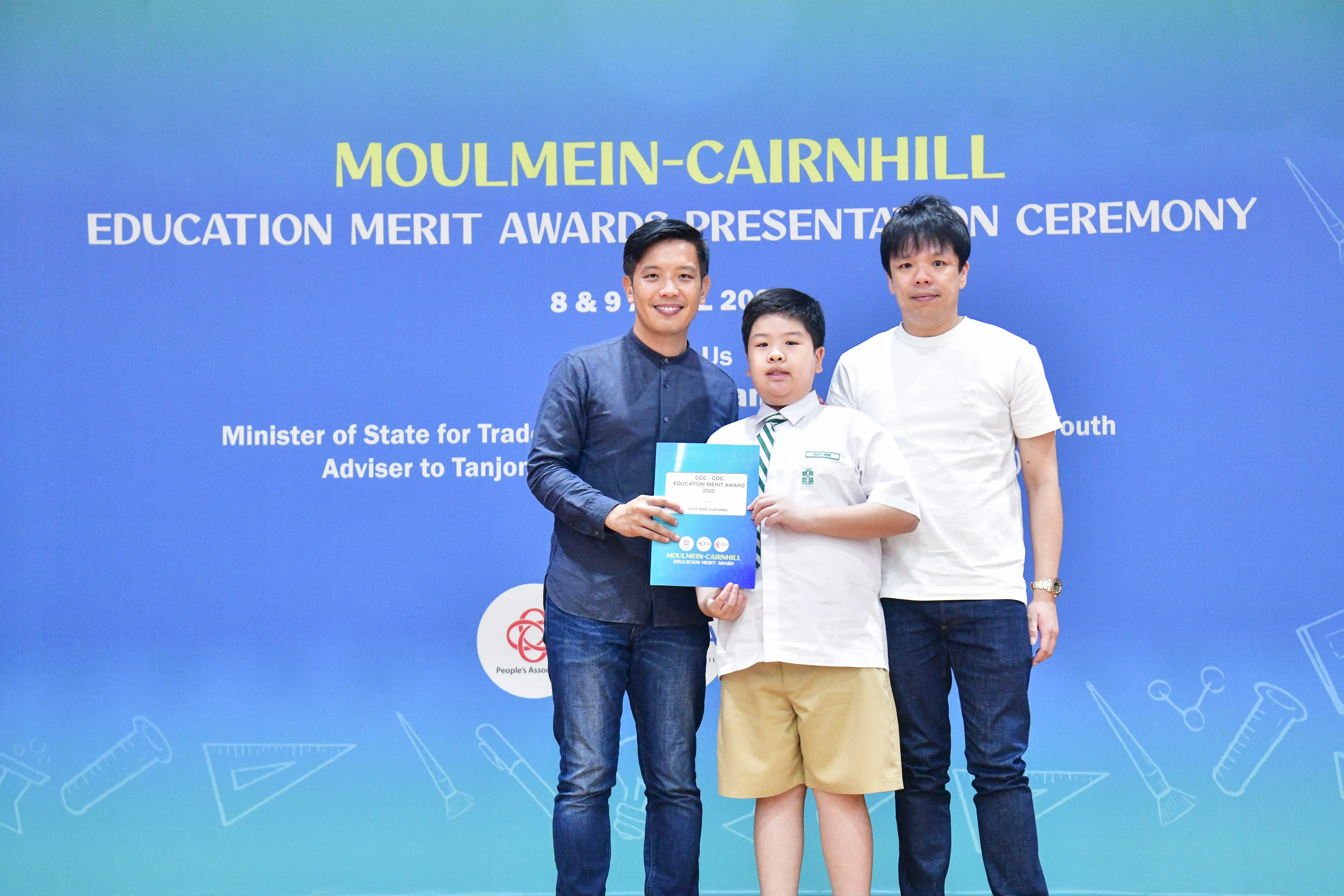 Two men and a boy in school uniform hold a certificate at the Moulmein-Cairnhill Education Merit Awards ceremony.