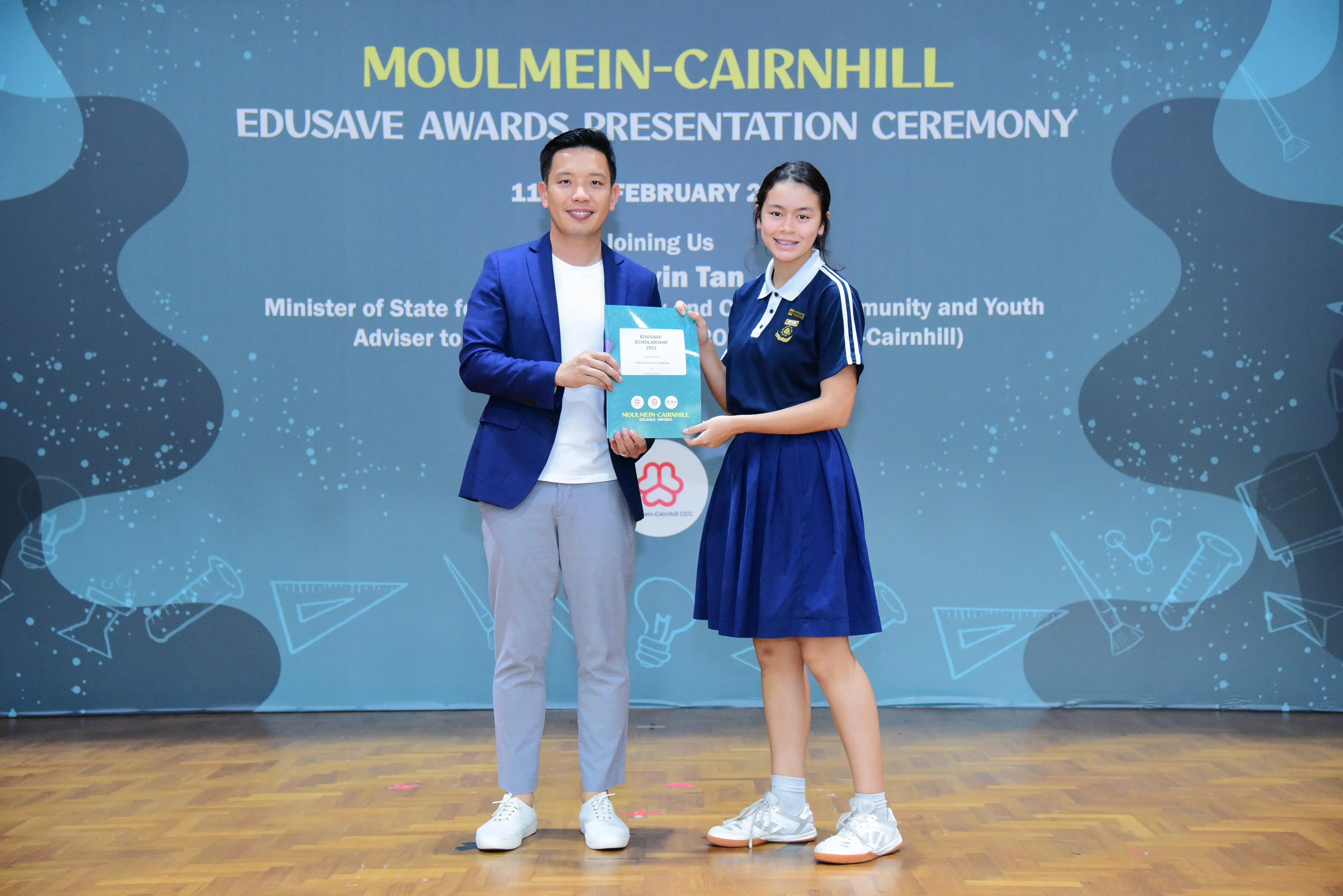 A man in a blue blazer and a girl in school uniform hold a certificate at the Moulmein-Cairnhill Edusave Awards Ceremony.