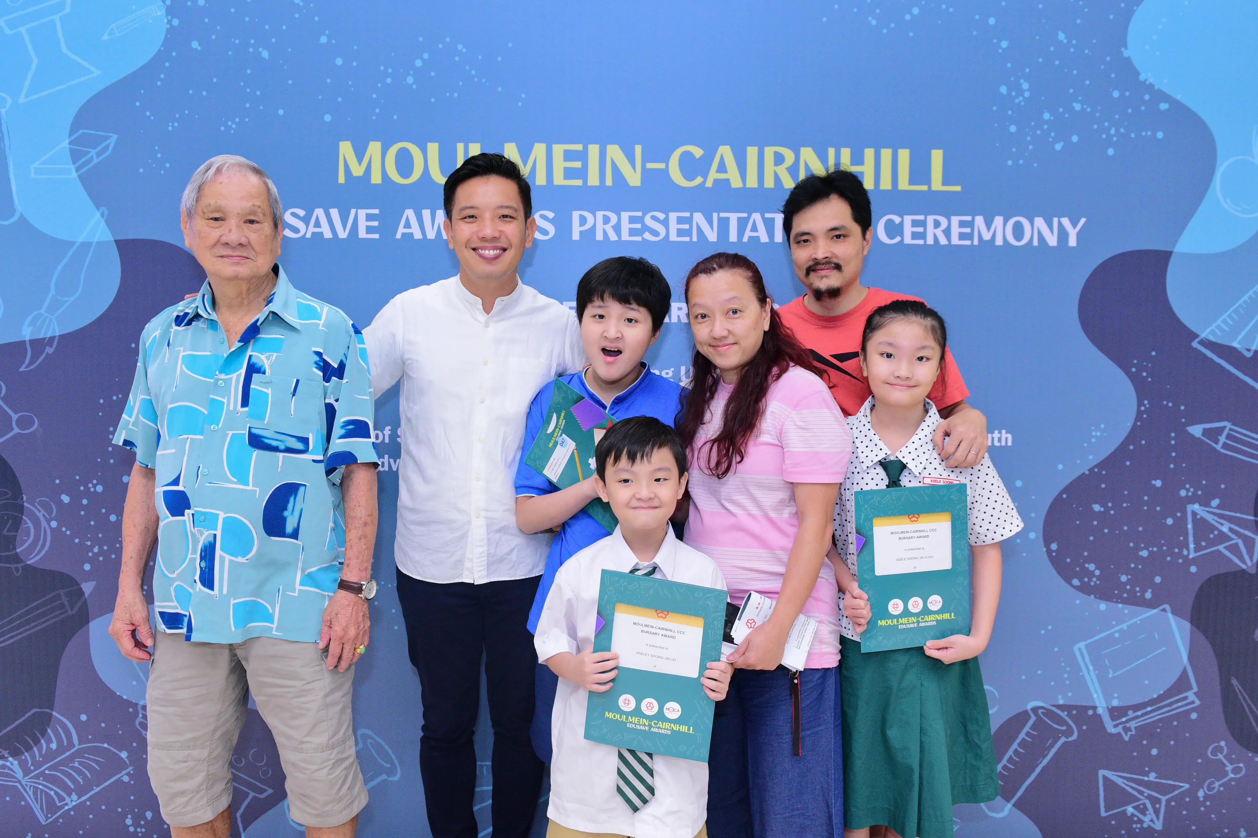 A family of seven poses at the Moulmein-Cairnhill Bursary Awards Presentation Ceremony, children holding award certificates.