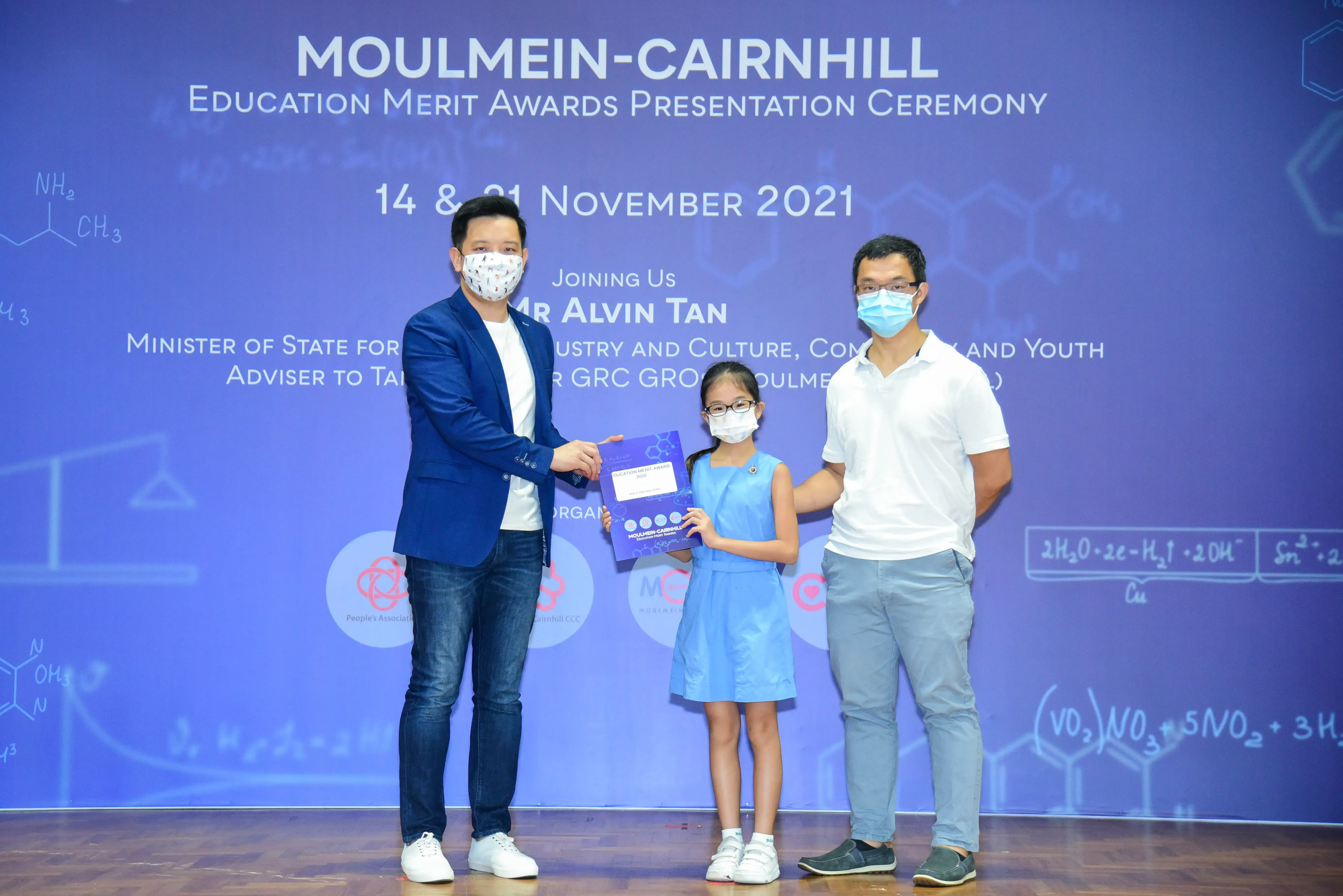 Two masked men and a young girl holding a certificate at the Moulmein-Cairnhill Education Merit Awards 2021.