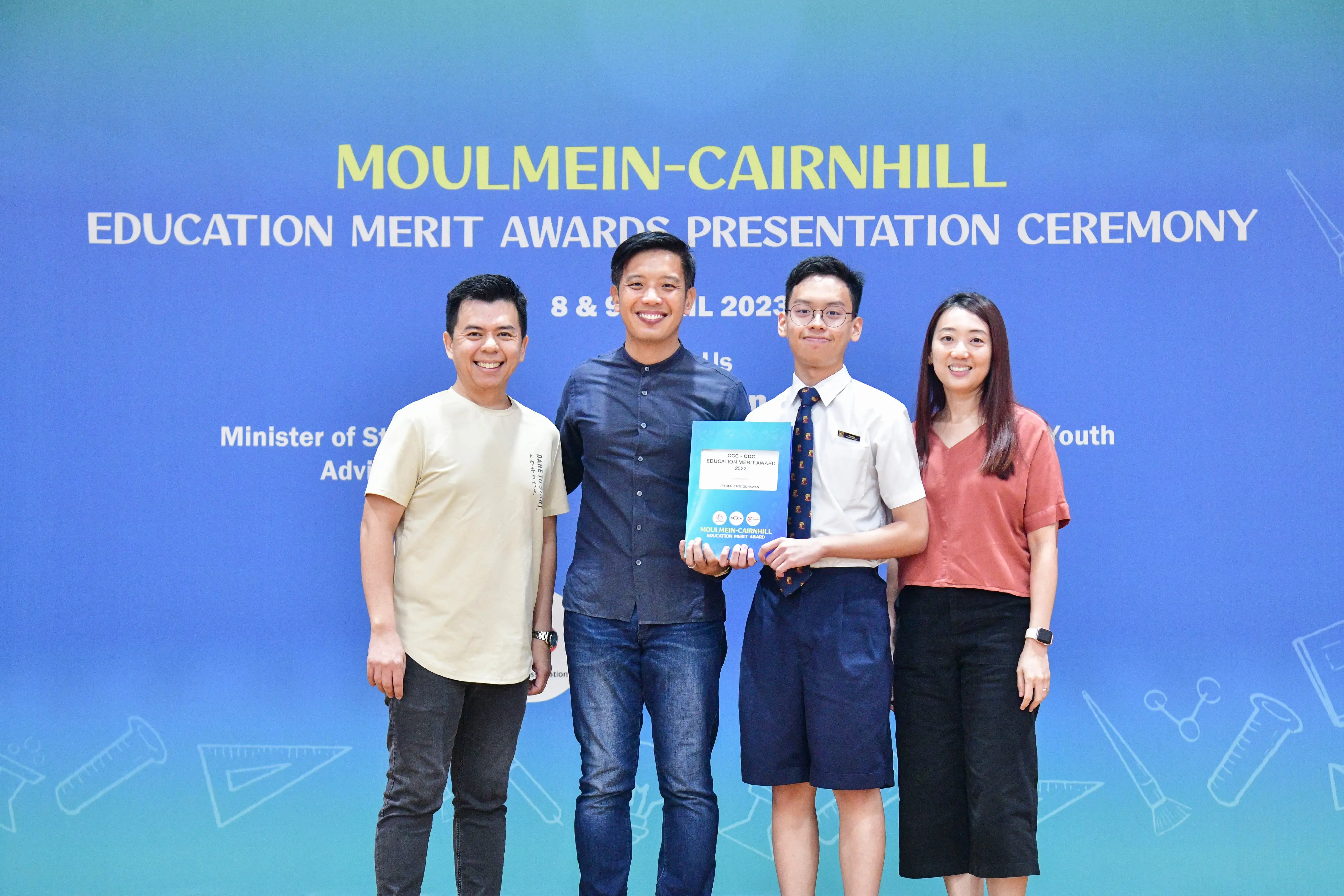 Four people stand smiling before a blue Moulmein-Cairnhill Education Merit Awards Presentation Ceremony backdrop, holding a certificate.