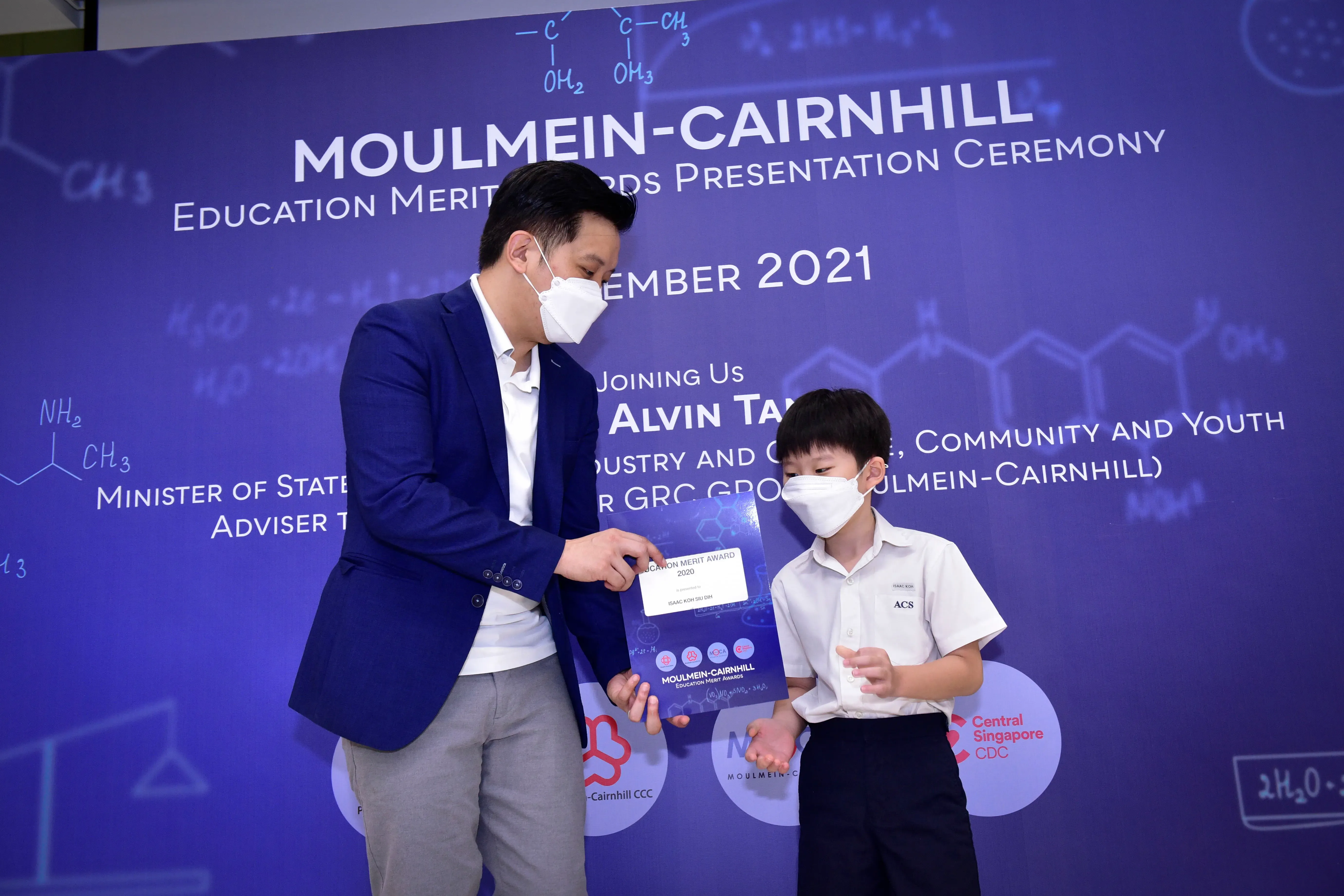 An adult in a blue blazer presents a certificate to a young boy in school uniform, both wearing masks, at a 2021 awards ceremony.
