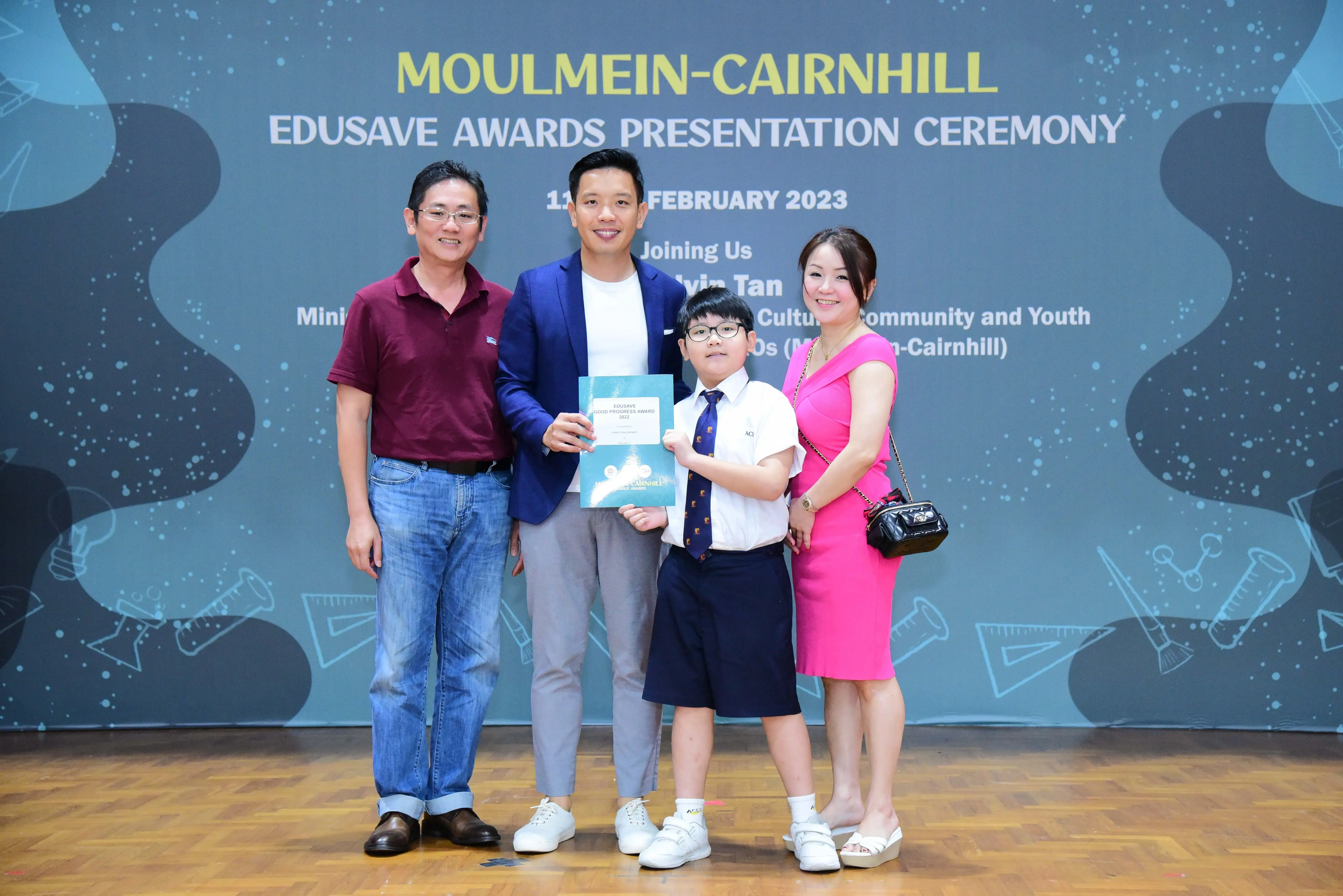 Four people pose at the Moulmein-Cairnhill Edusave Awards Presentation Ceremony, with a boy holding a certificate.