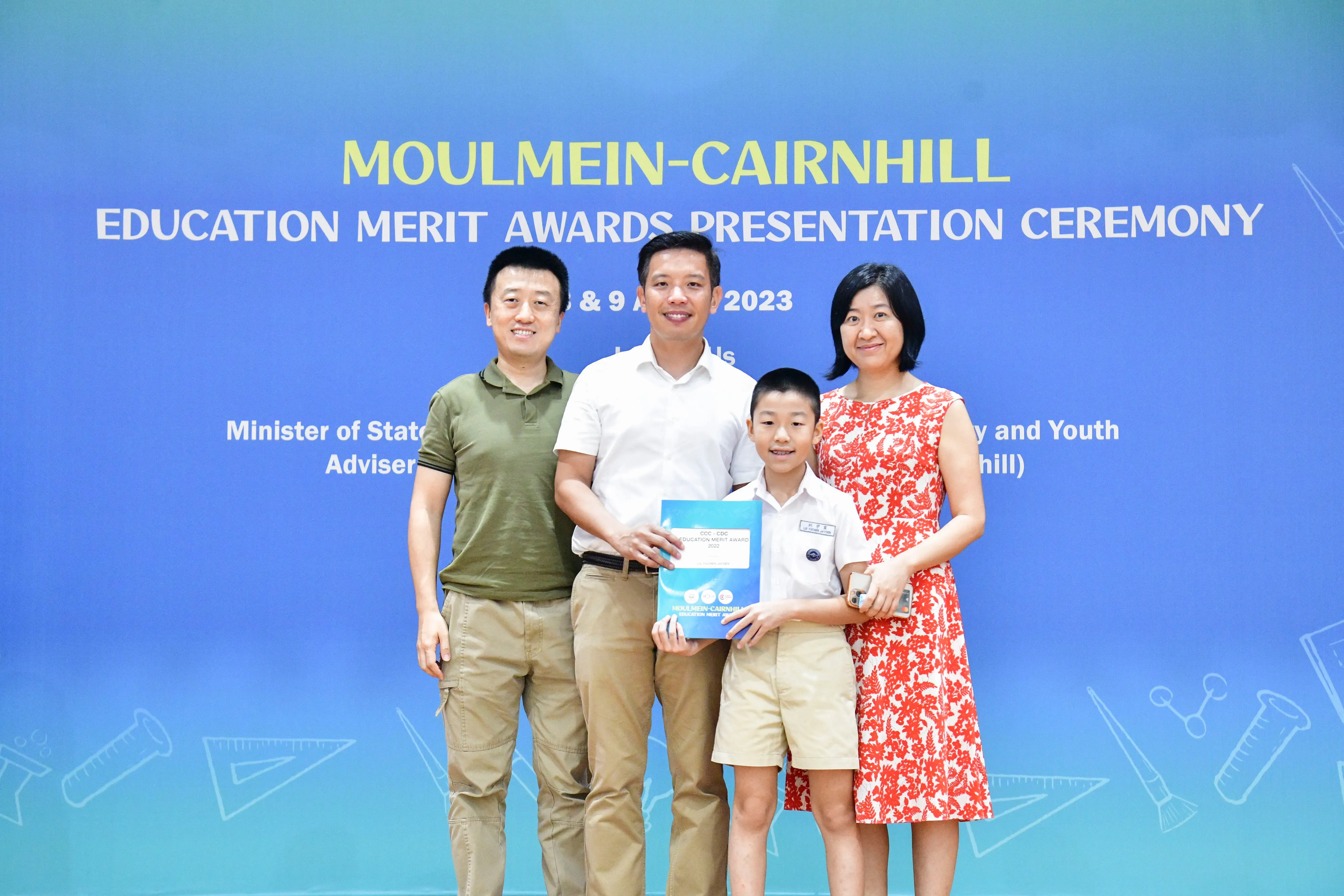 Three adults and a boy holding an award folder pose in front of a Moulmein-Cairnhill Education Merit Awards 2023 banner.