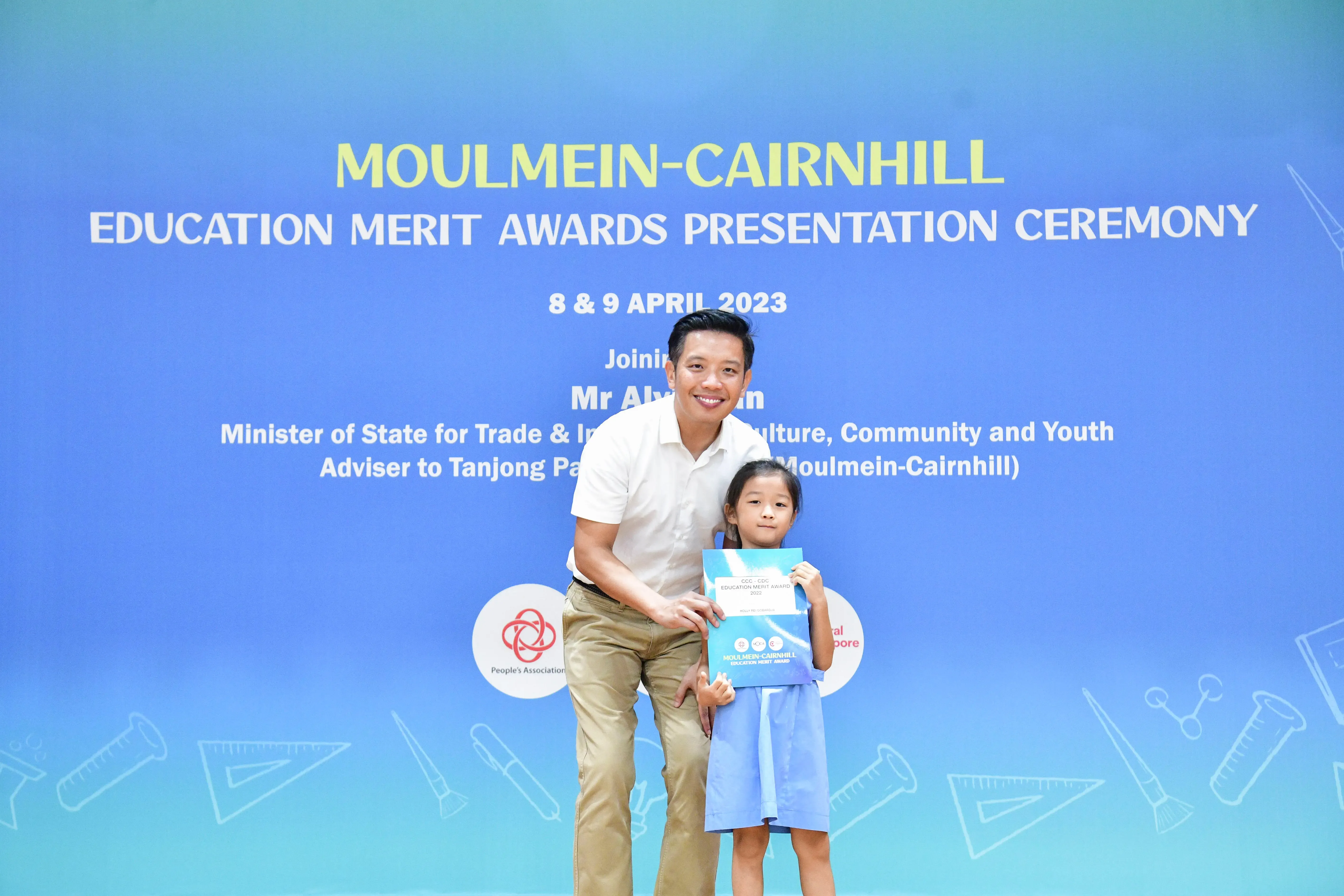 A man and a young girl pose with an award in front of a Moulmein-Cairnhill Education Merit Awards banner.