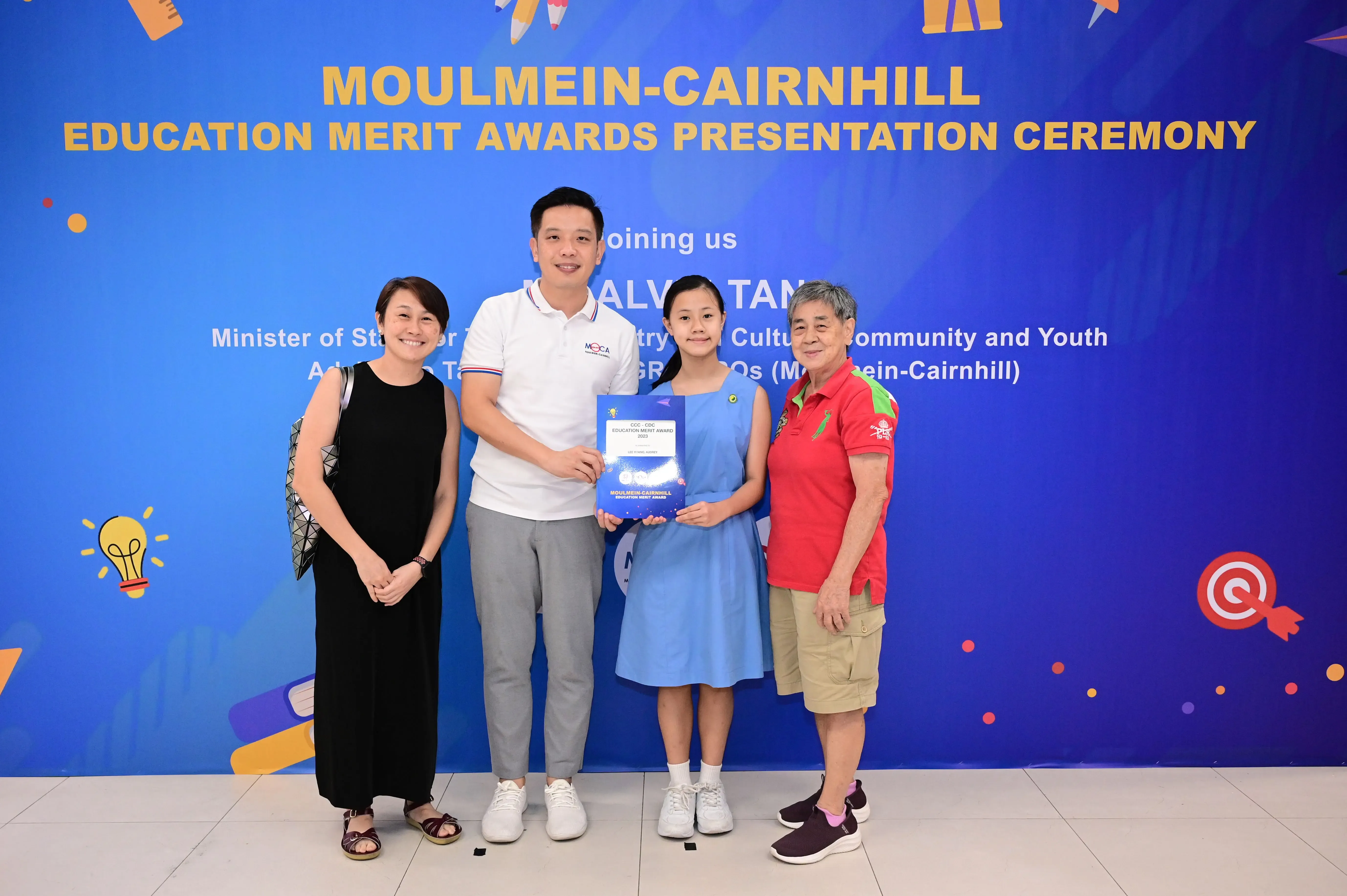 Four people pose in front of a Moulmein-Cairnhill Education Merit Awards Presentation Ceremony banner, girl holds award.