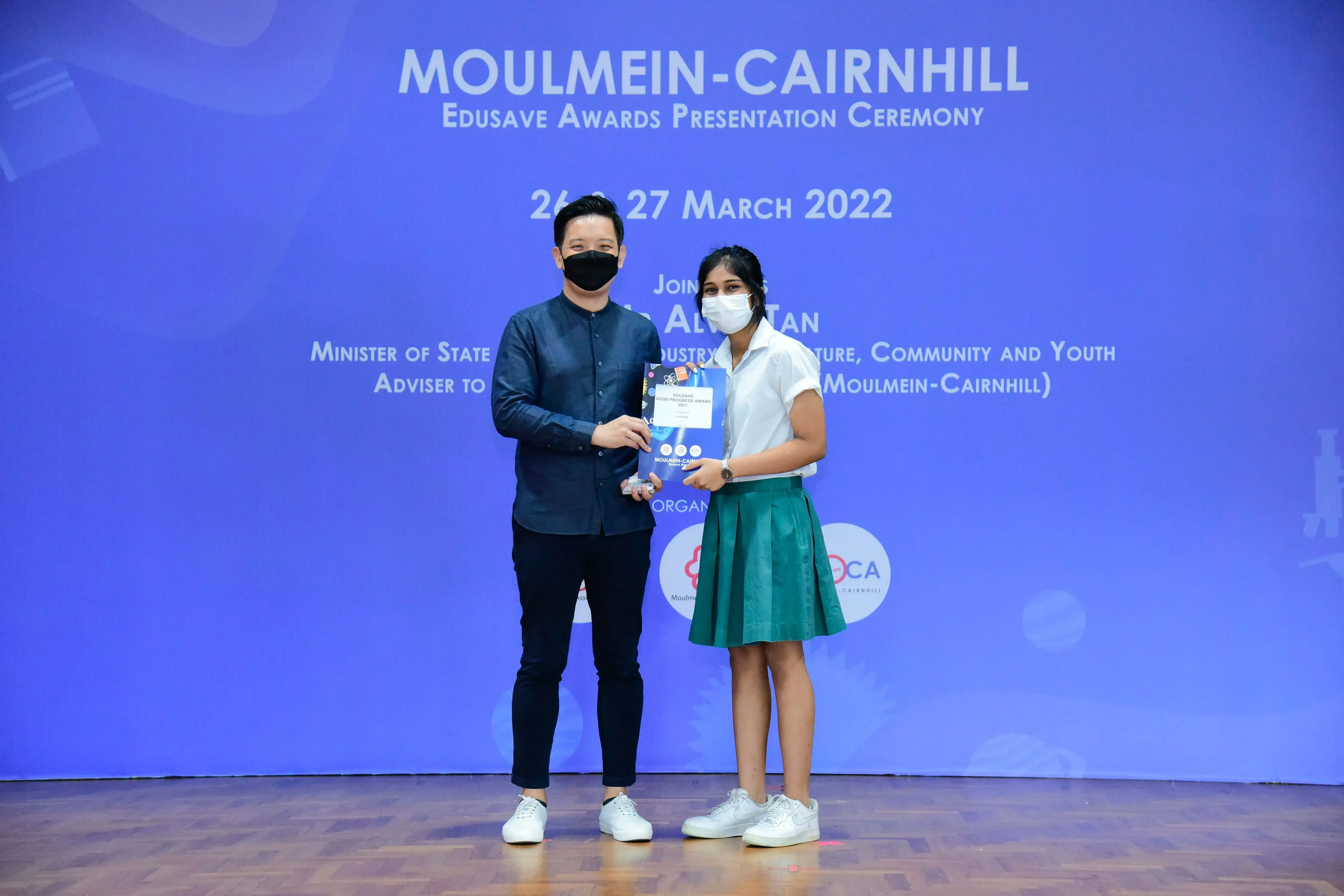 Two masked people hold an award at the Moulmein-Cairnhill Edusave Awards Presentation Ceremony, March 2022.