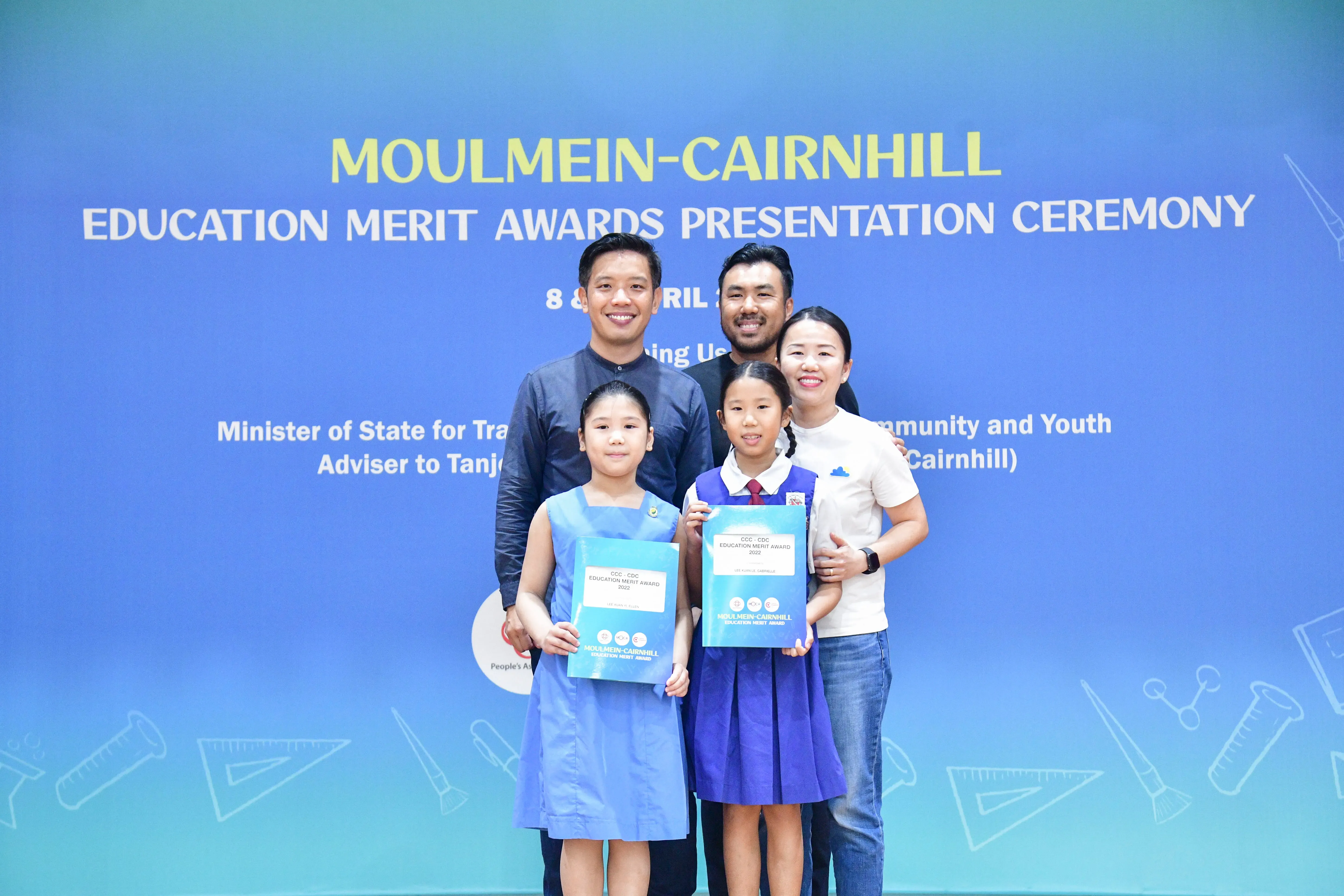 Two adults and two girls holding award certificates pose before a Moulmein-Cairnhill Education Merit Awards banner.
