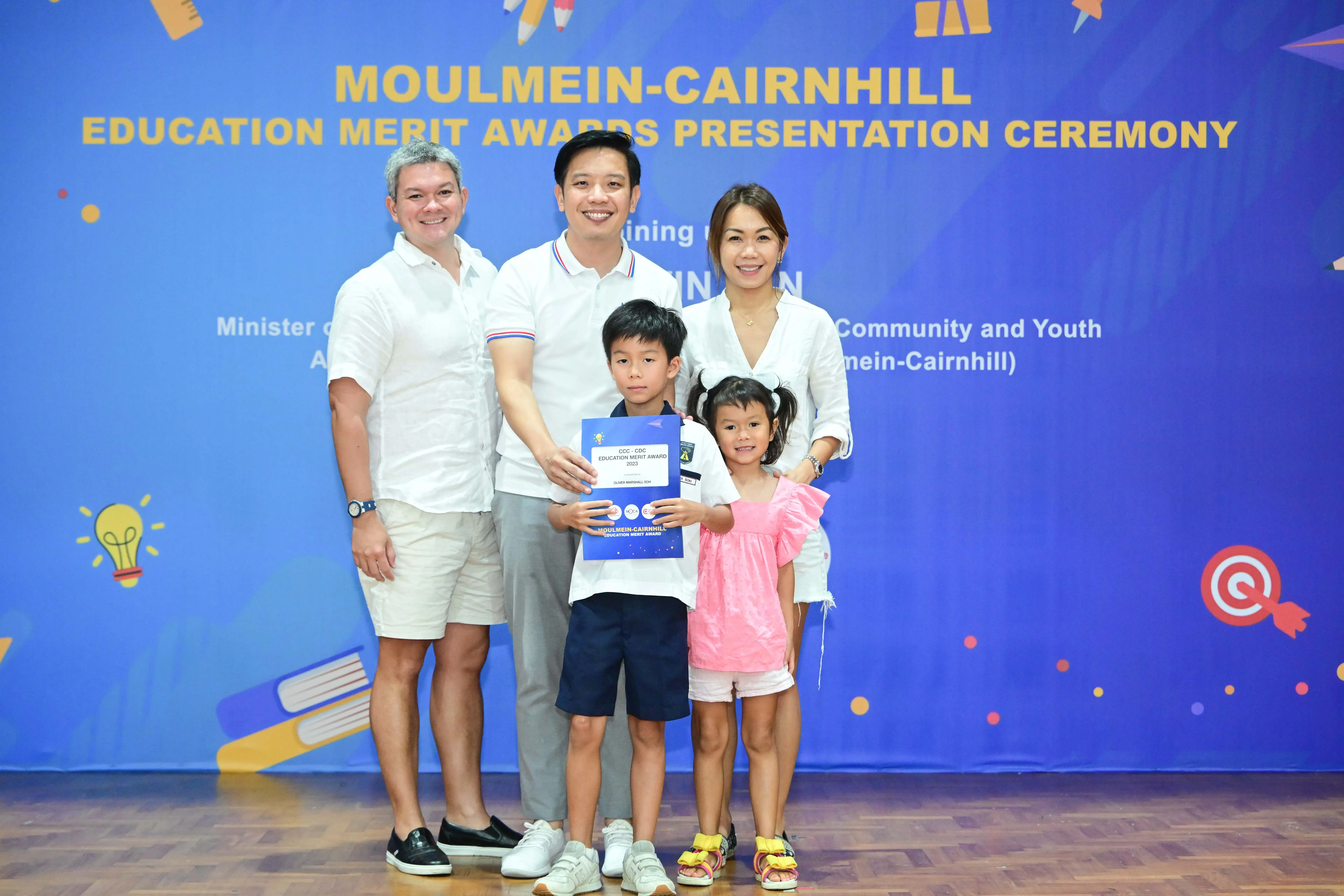 Family of five posing with a merit award certificate in front of a Moulmein-Cairnhill Education Merit Awards banner.
