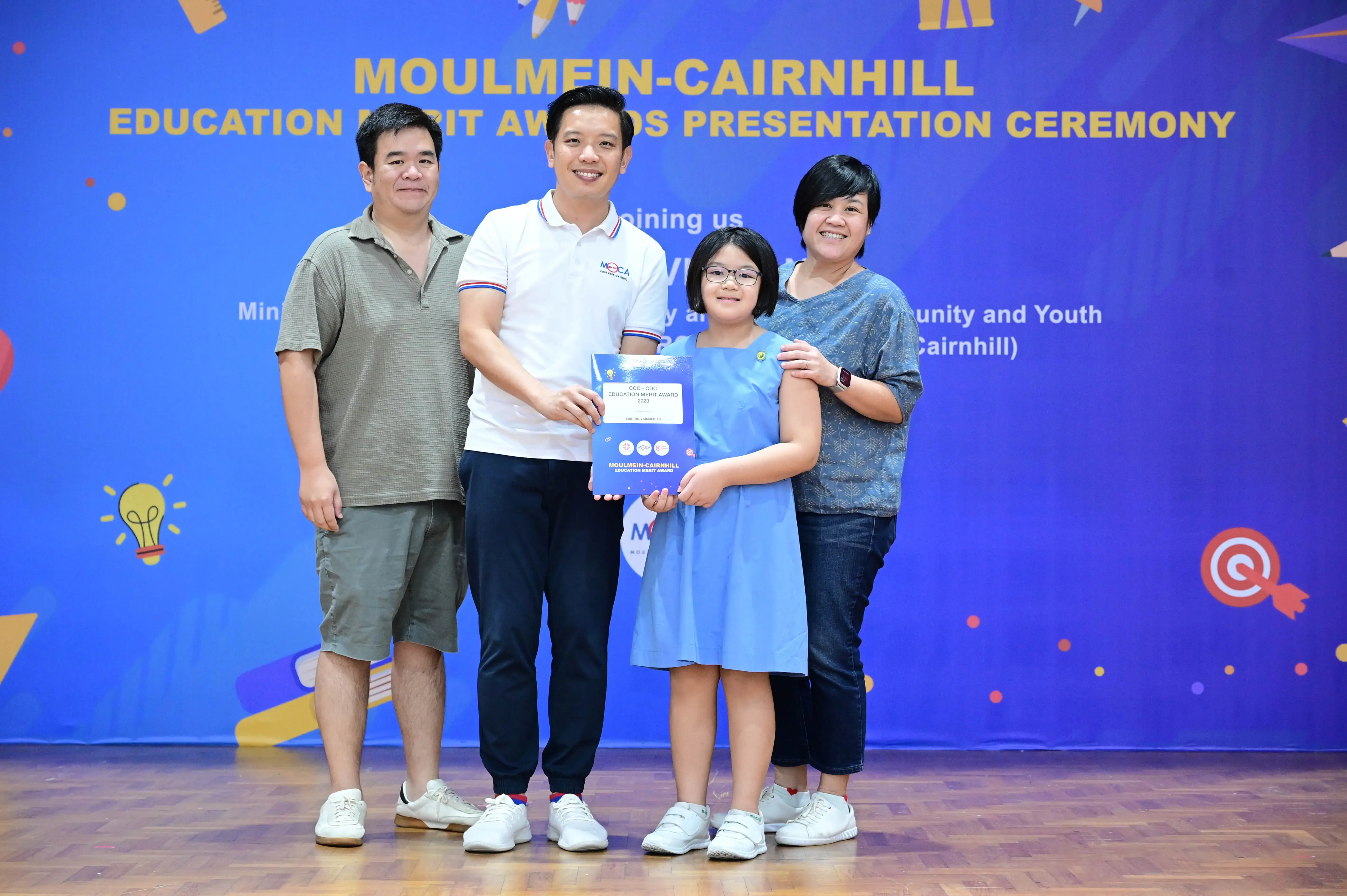 A girl holding an award certificate poses with three adults at the Moulmein-Cairnhill Education Merit Awards ceremony.