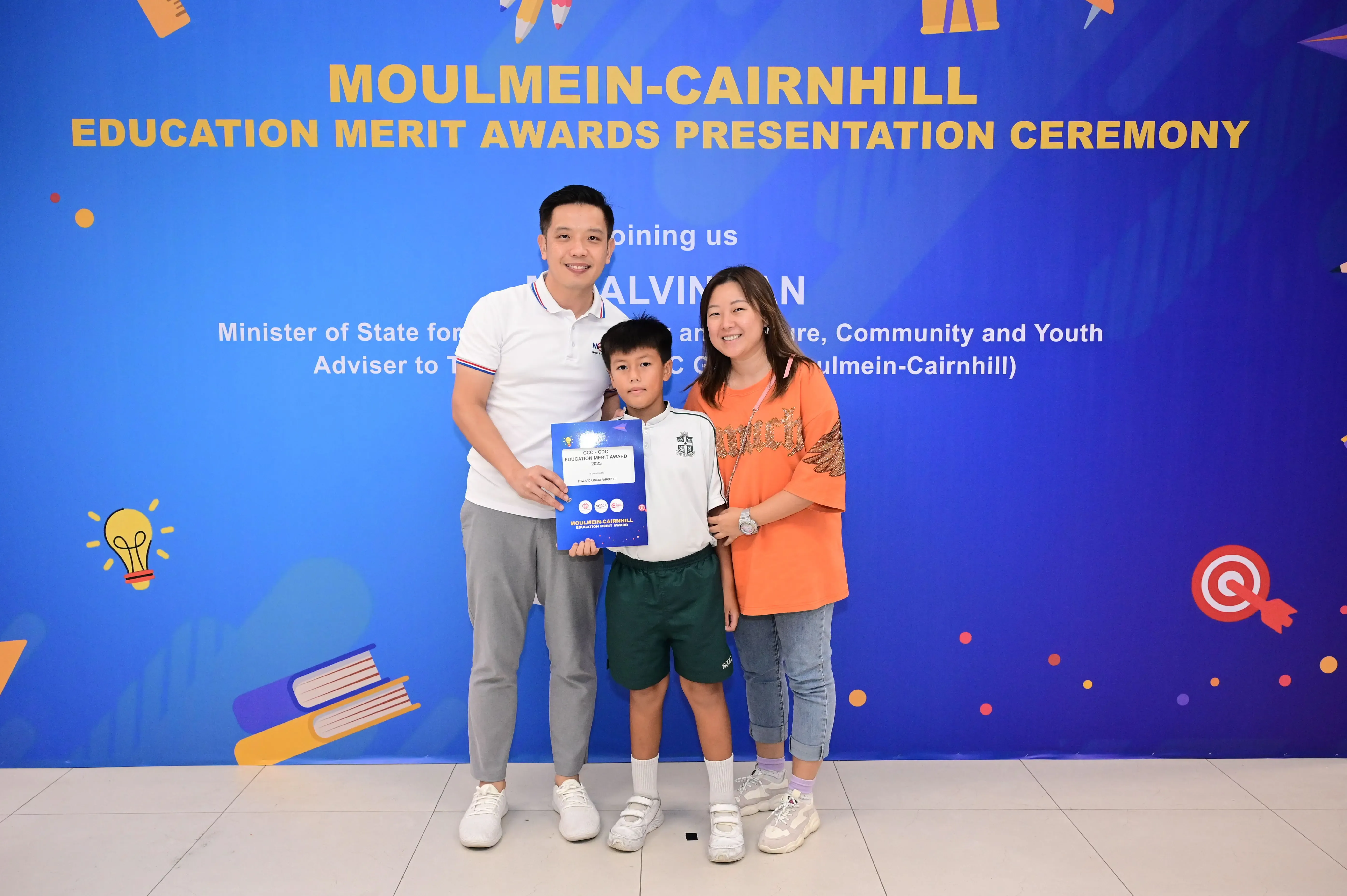 A boy holding an award certificate stands between two adults in front of a Moulmein-Cairnhill Education Merit Awards banner.