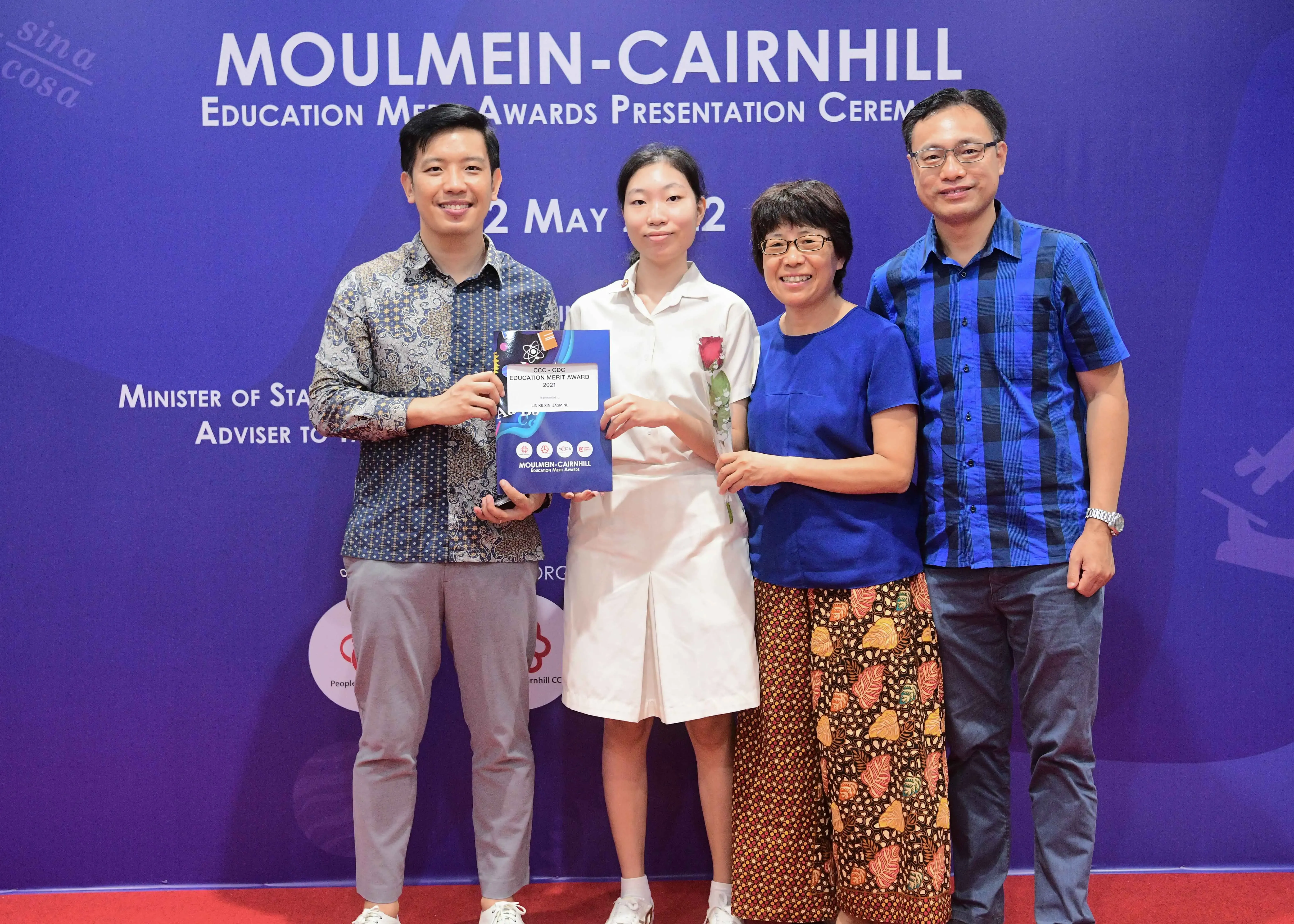 Four people pose at the Moulmein-Cairnhill Education Merit Awards Presentation Ceremony, holding a certificate and a rose.