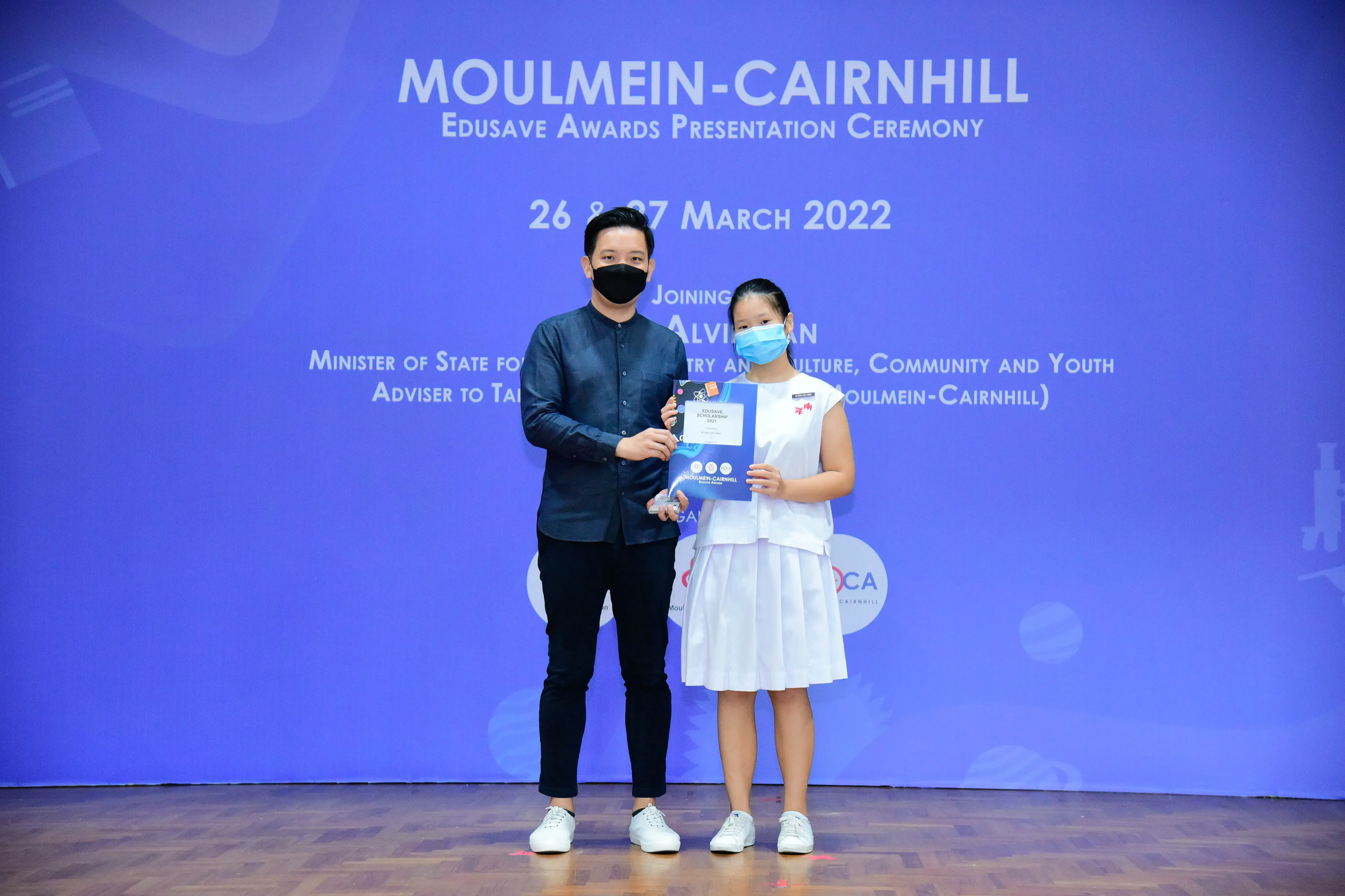 A masked man and young girl hold an award on stage at the Moulmein-Cairnhill Edusave Awards Ceremony, March 2022.