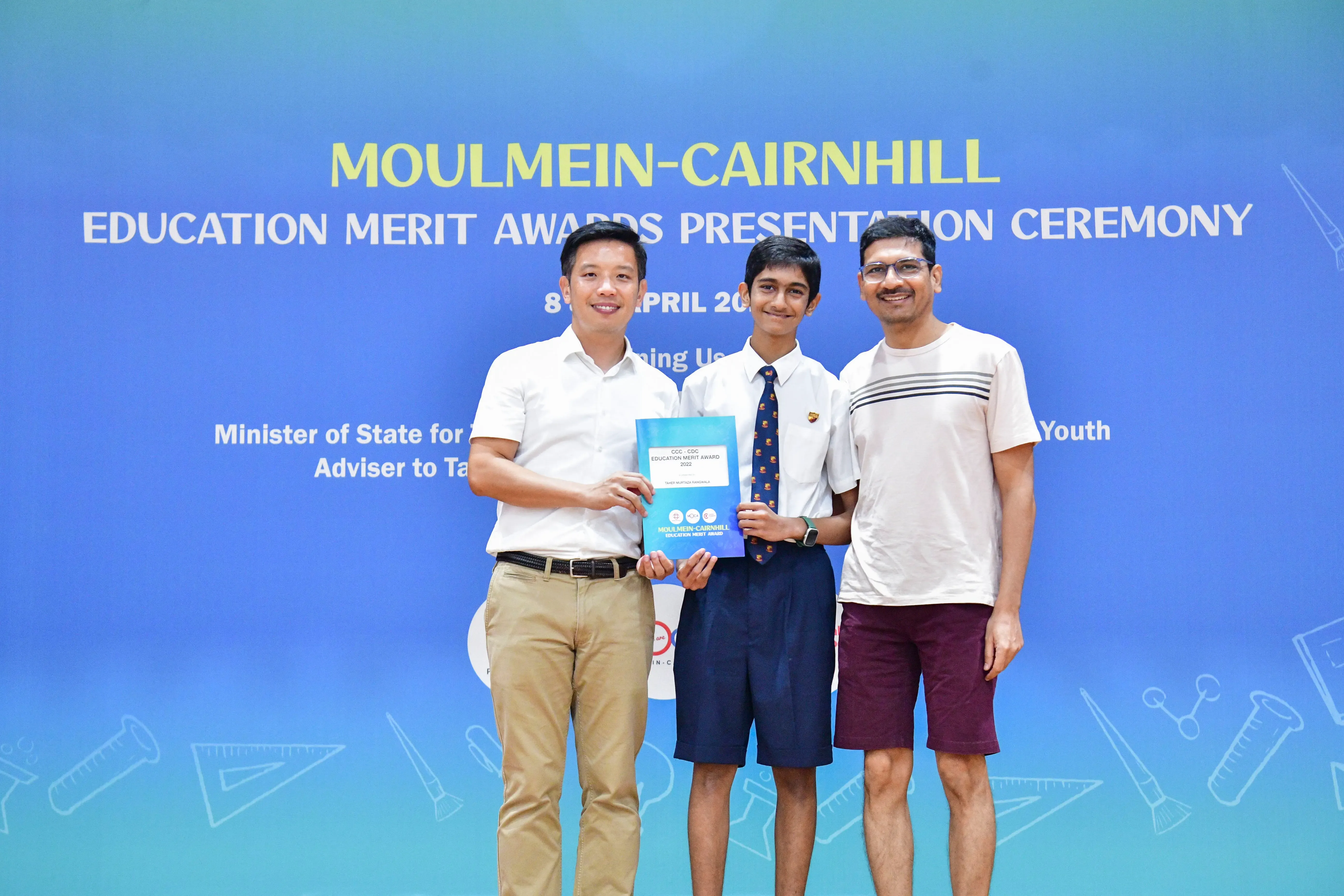 Three smiling people stand before a blue Moulmein-Cairnhill Education Merit Awards banner, with a student holding a certificate.