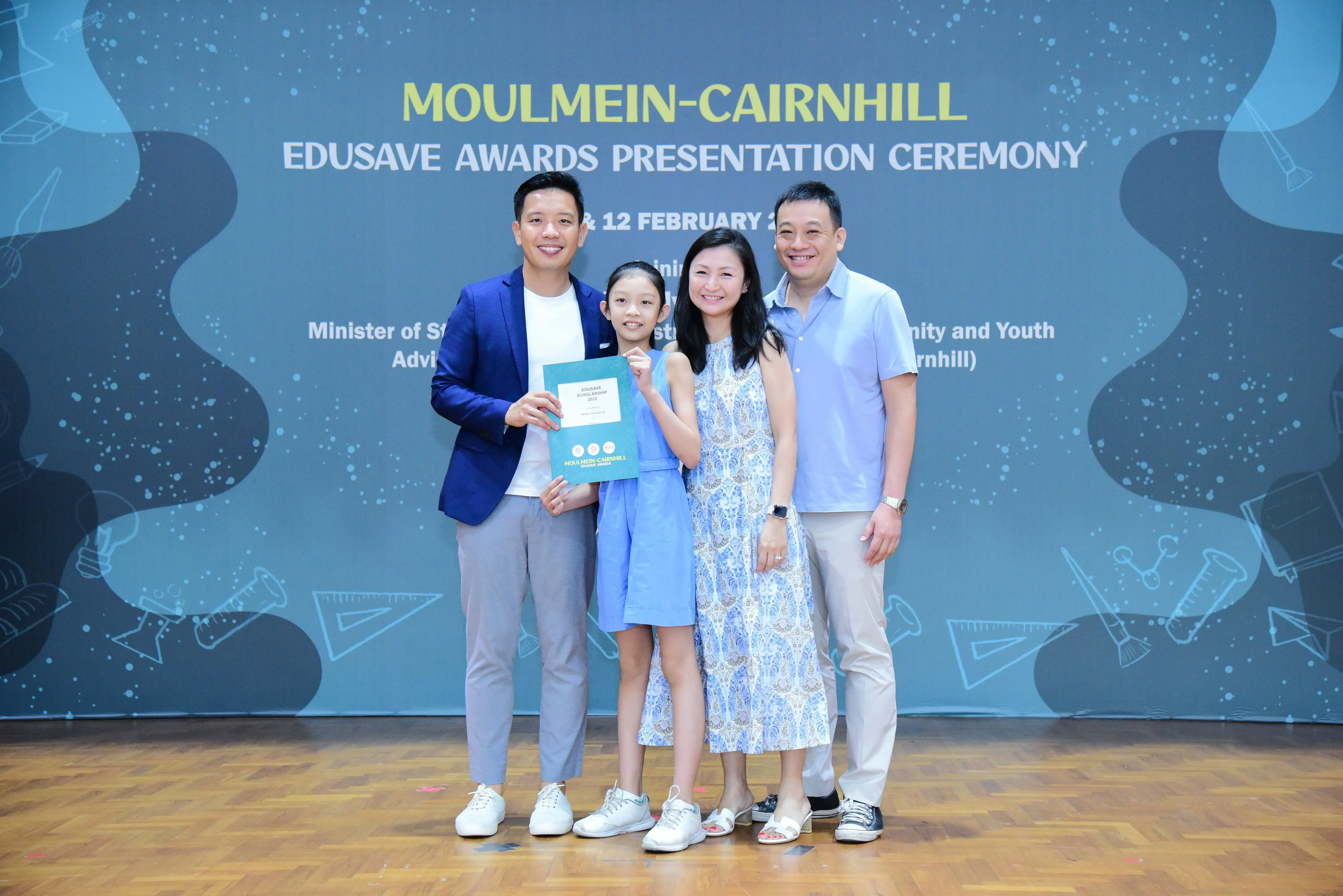 Four people smile holding a certificate at the Moulmein-Cairnhill Edusave Awards Presentation Ceremony backdrop.