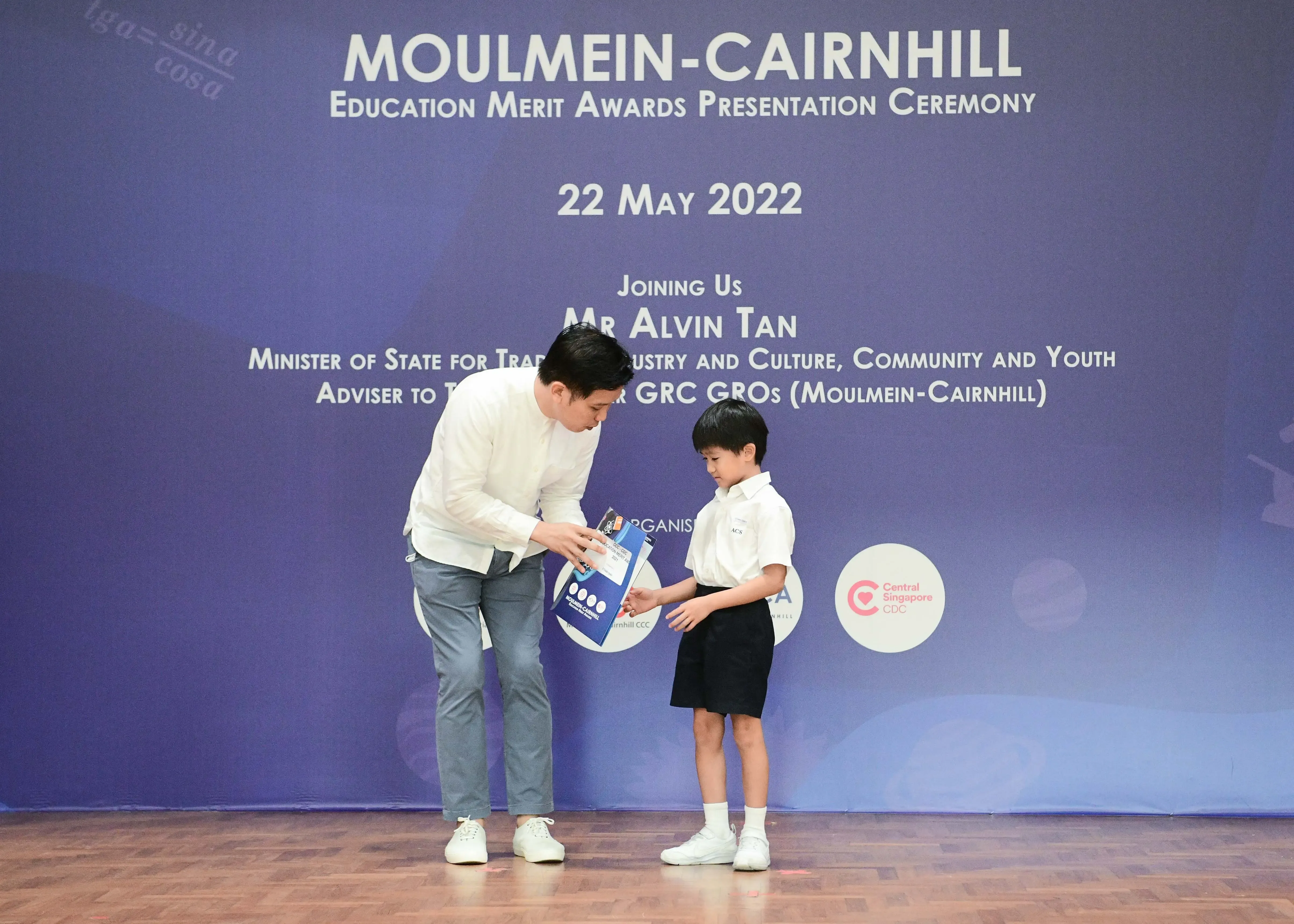 An adult in white shirt hands an award to a young student on stage at the Moulmein-Cairnhill Education Merit Awards 2022.