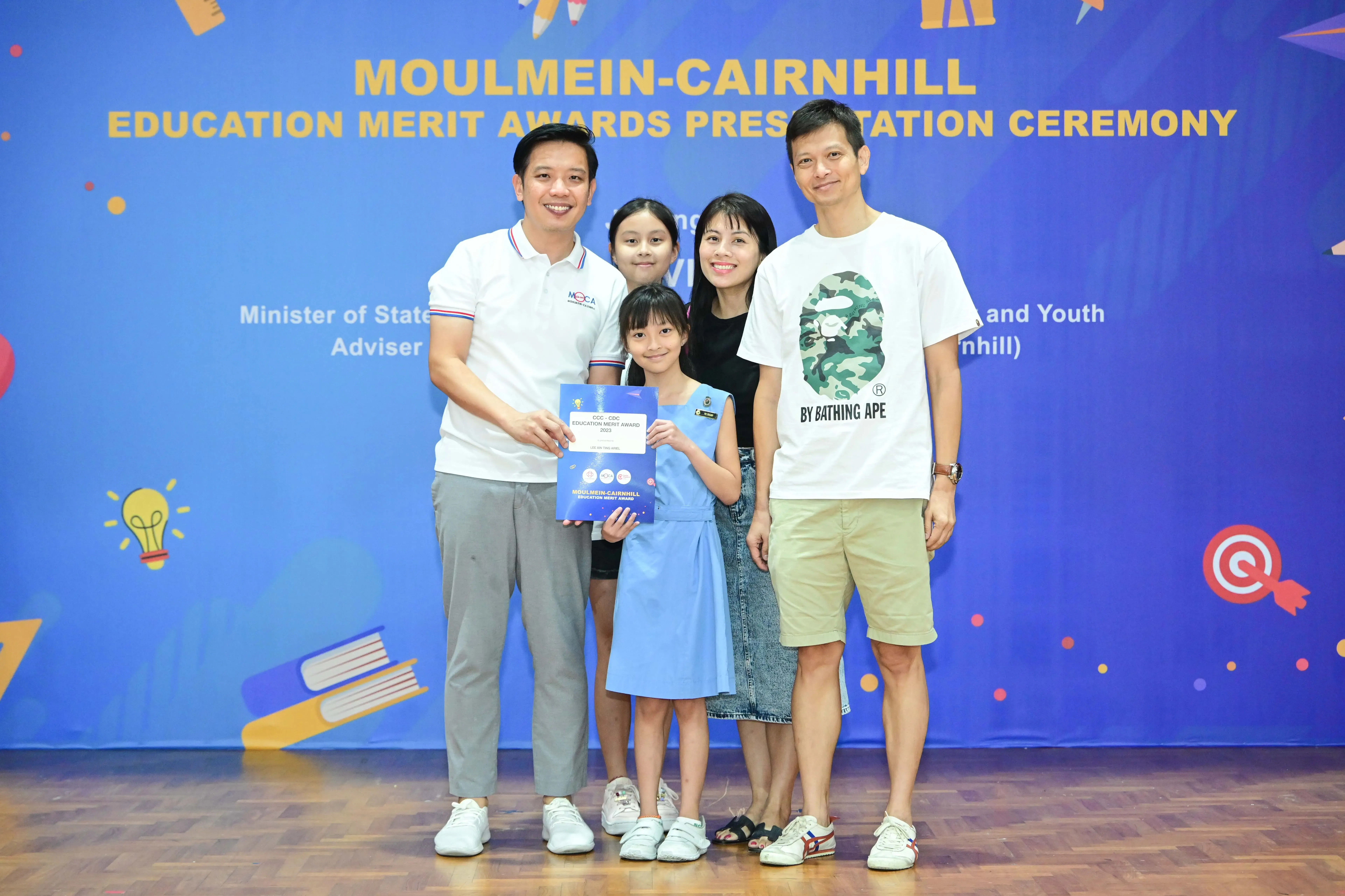 A girl in a blue dress holds an award certificate with four adults at the Moulmein-Cairnhill Education Merit Awards ceremony.