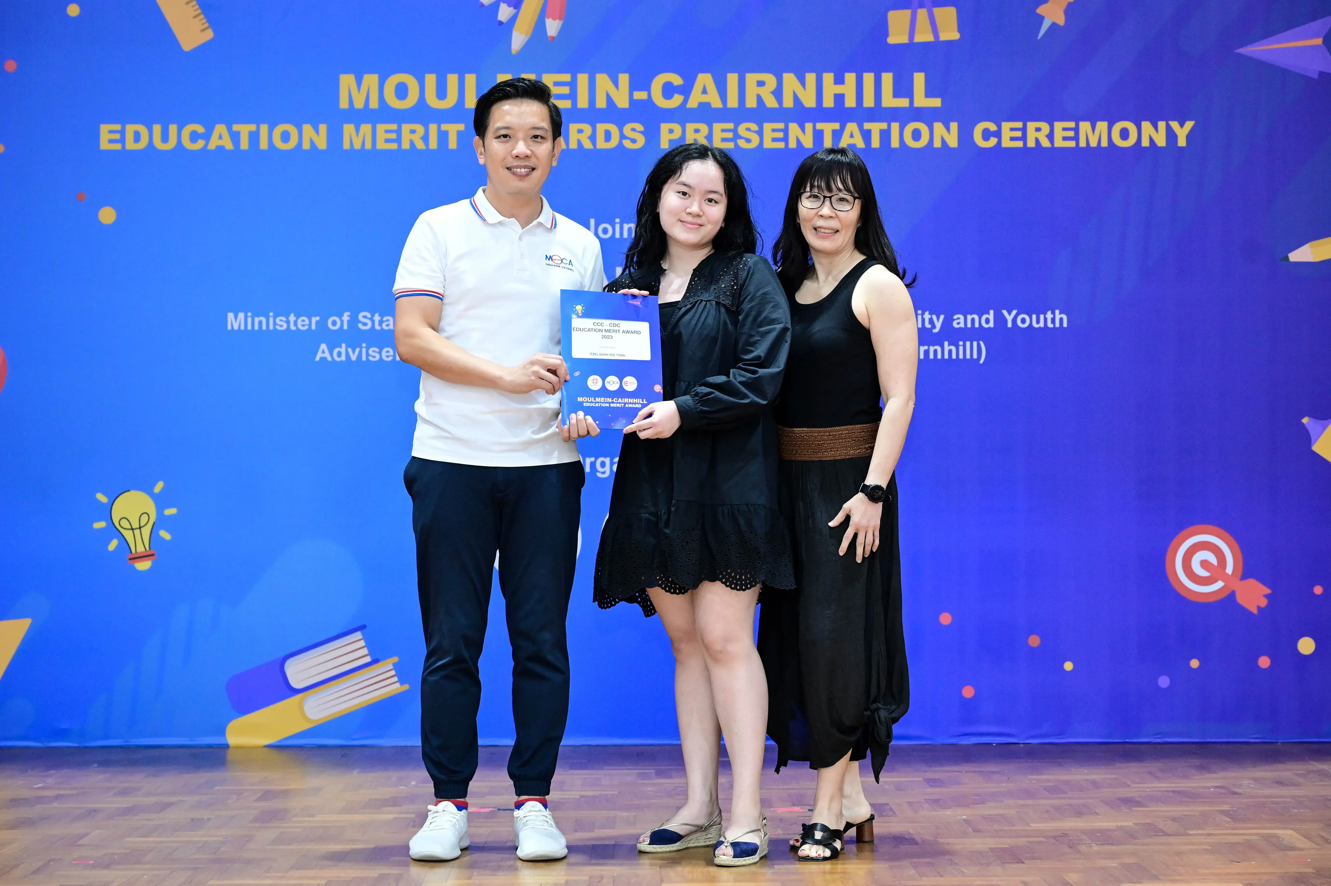 Three people pose together holding an award certificate at the Moulmein-Cairnhill Education Merit Awards Presentation Ceremony.