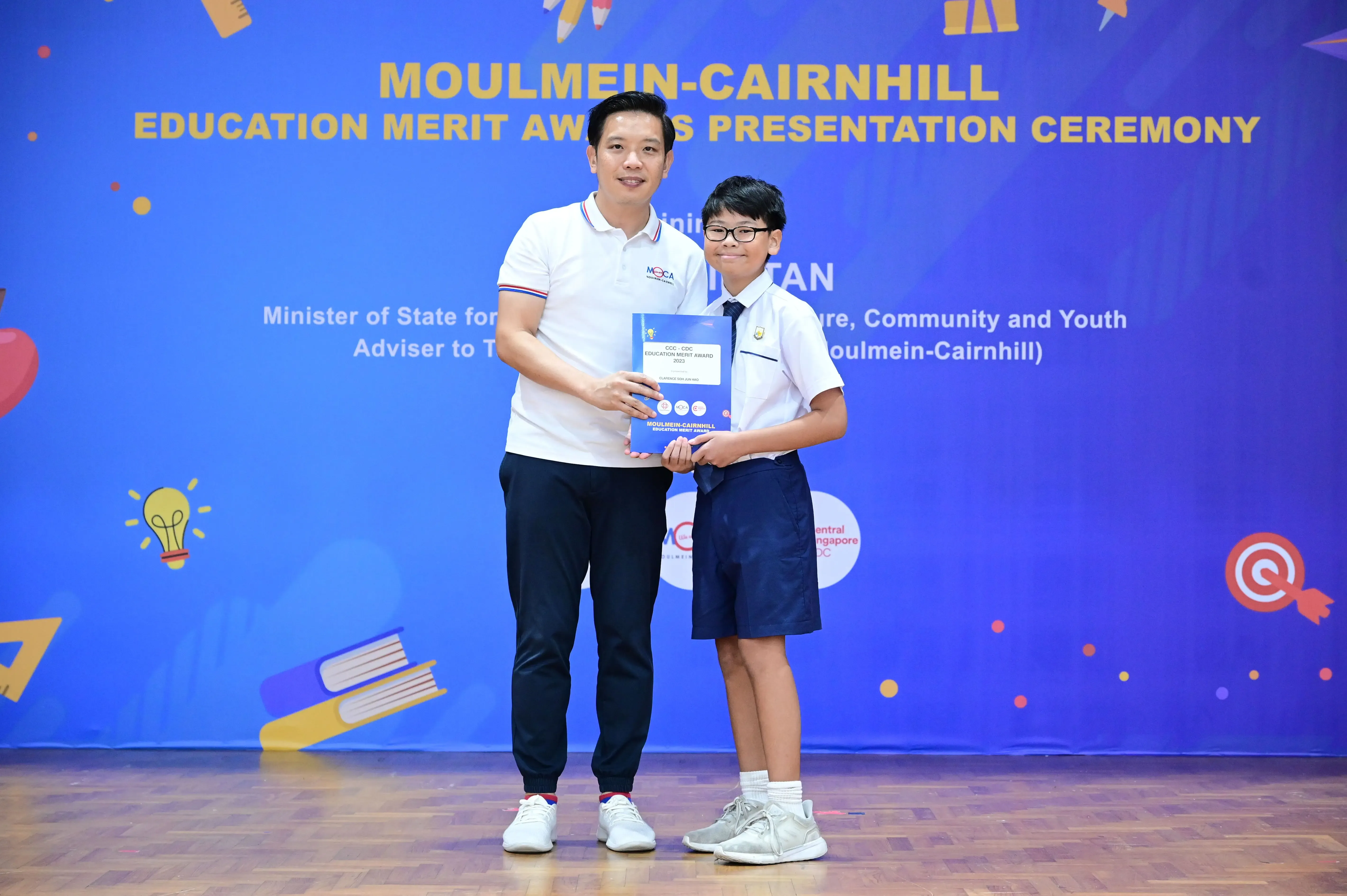 An adult and a young student hold a certificate together at the Moulmein-Cairnhill Education Merit Awards ceremony.