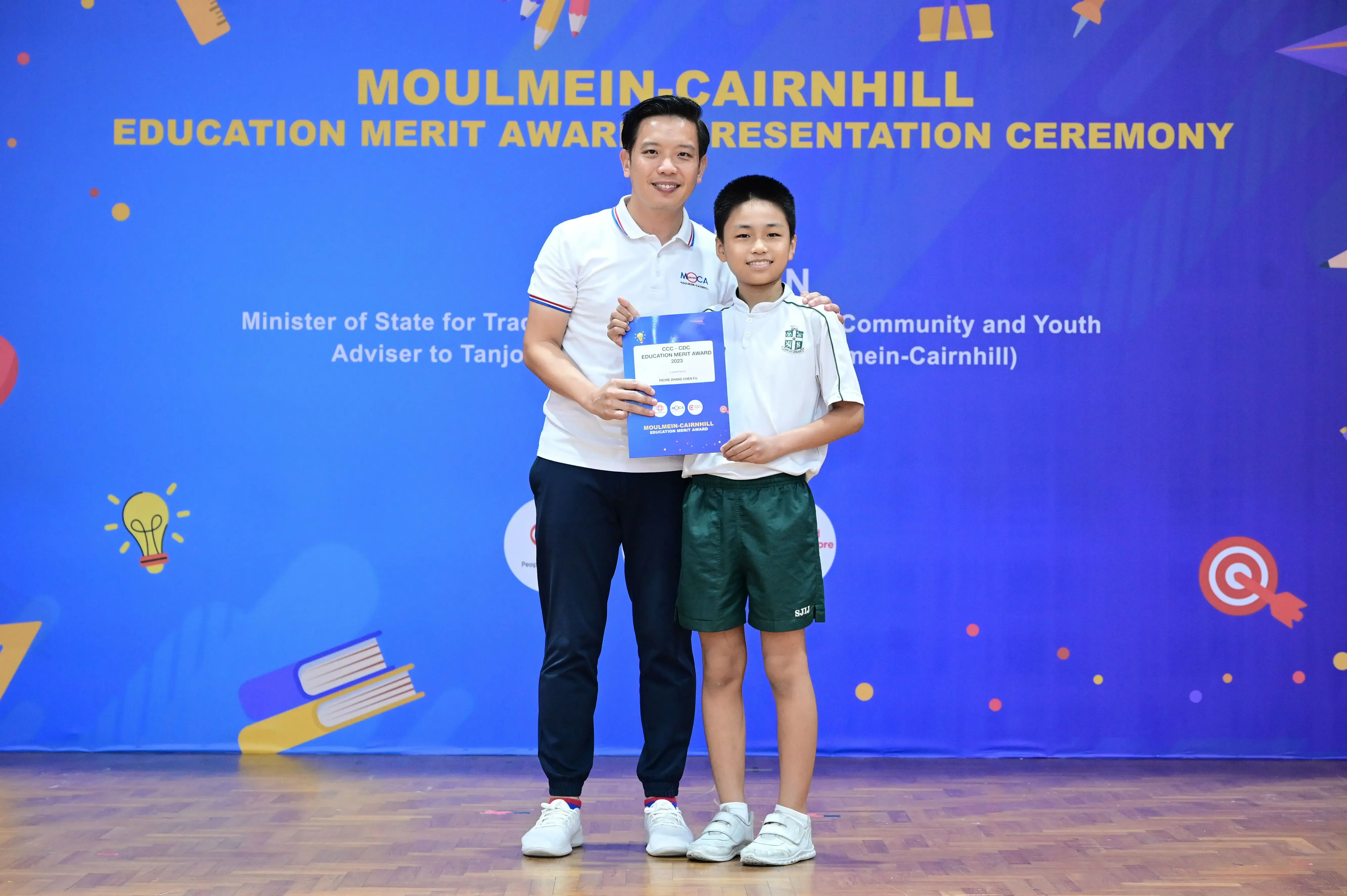 An adult and a young student hold an award certificate at the Moulmein-Cairnhill Education Merit Award Ceremony.