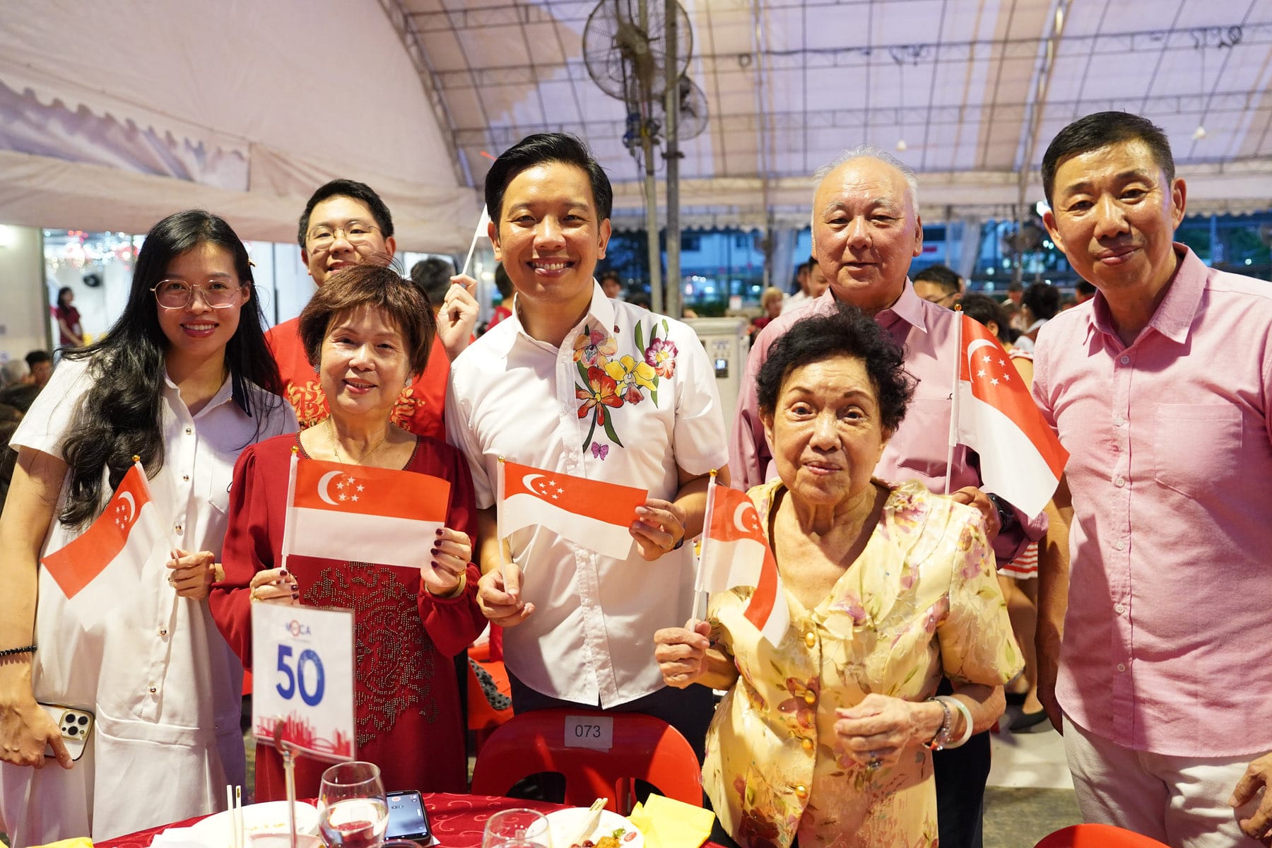 Group of people smiling and holding small Singapore flags at an outdoor celebration under a tent.