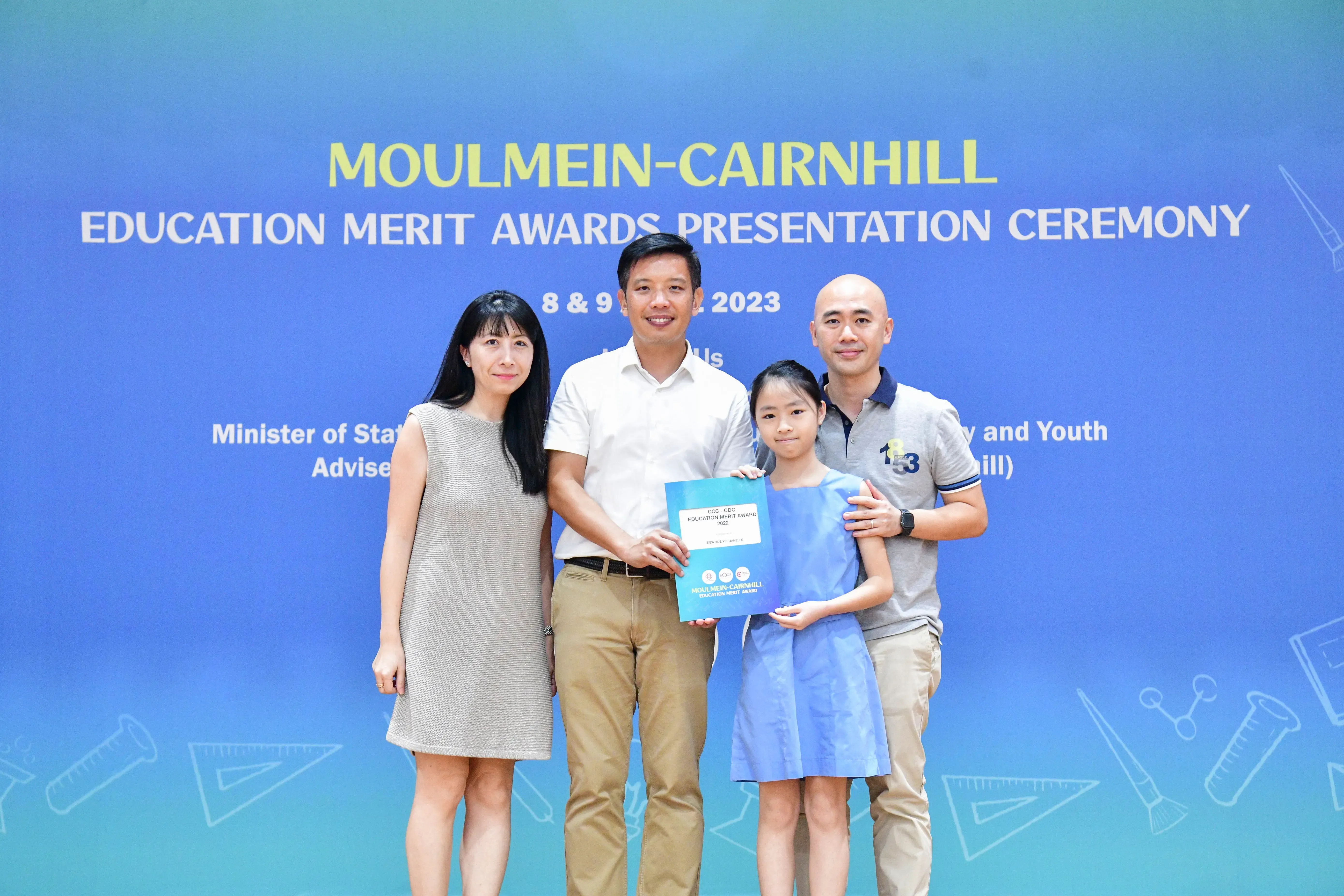 Four people pose before a blue Moulmein-Cairnhill Education Merit Awards 2023 banner, with a girl holding an award folder.