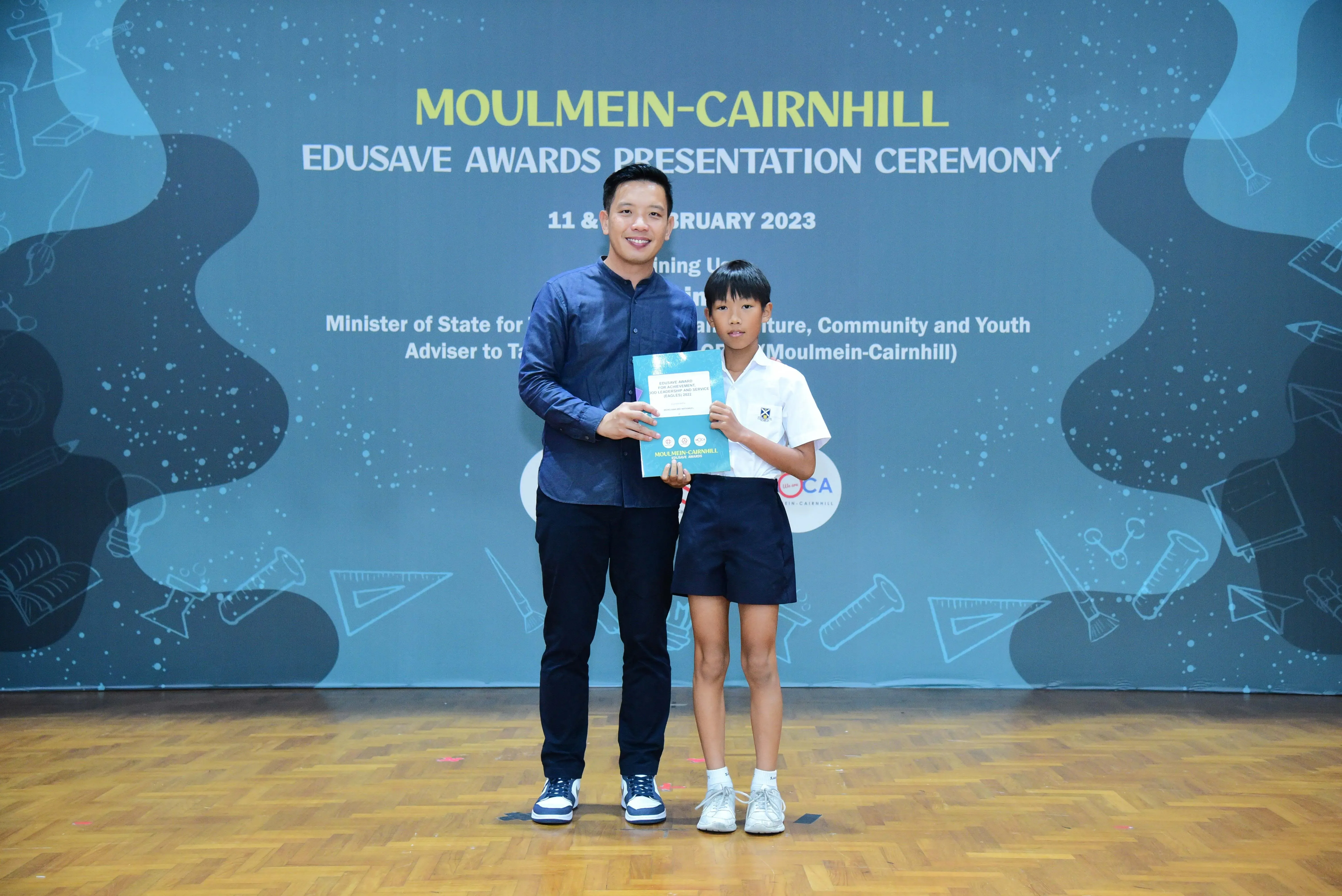 An adult and a young student hold a certificate together at the Moulmein-Cairnhill Edusave Awards Ceremony 2023.