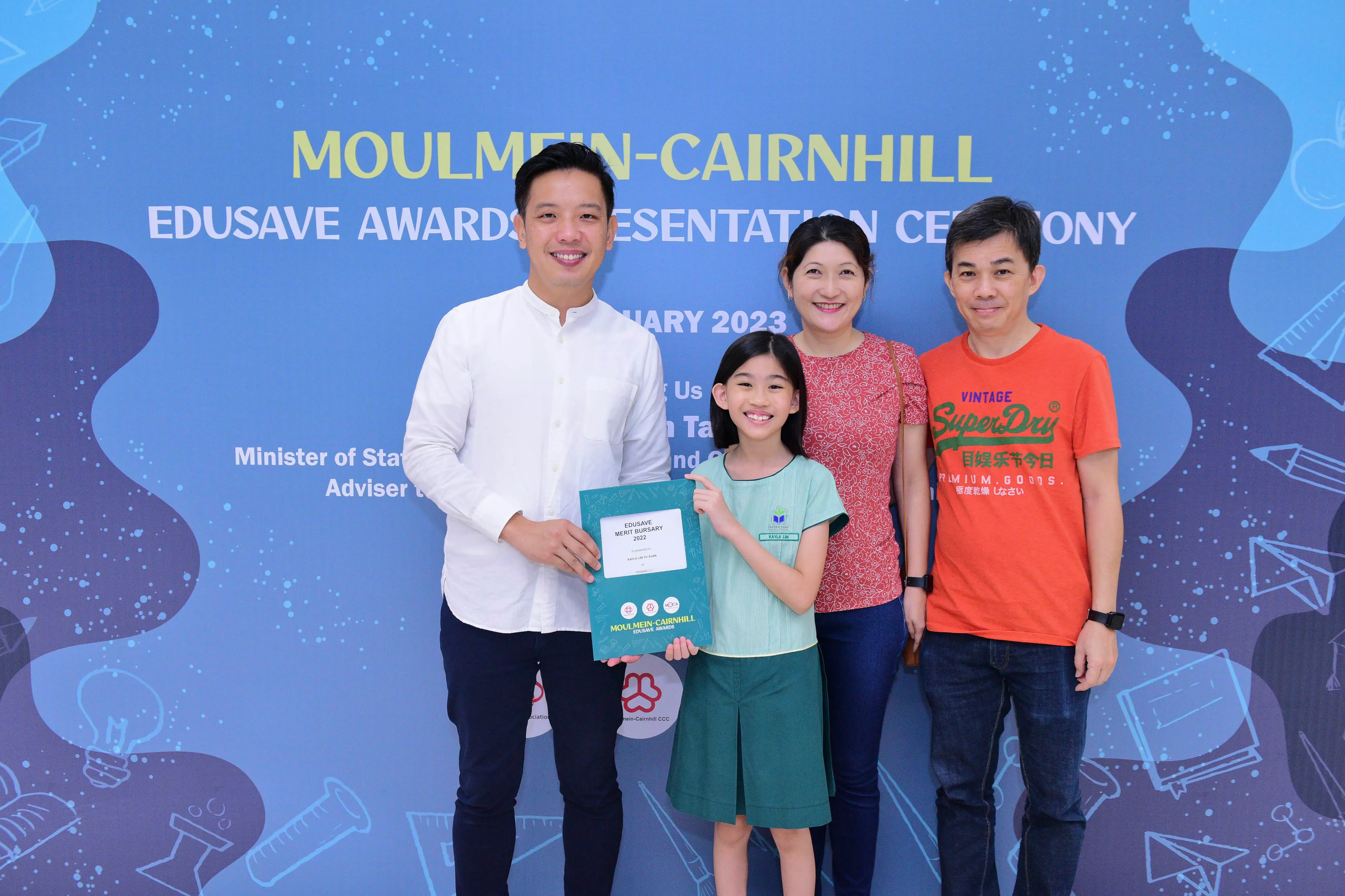 A girl holds an Edusave Merit Bursary certificate with three smiling adults at the Moulmein-Cairnhill Awards Ceremony 2023.