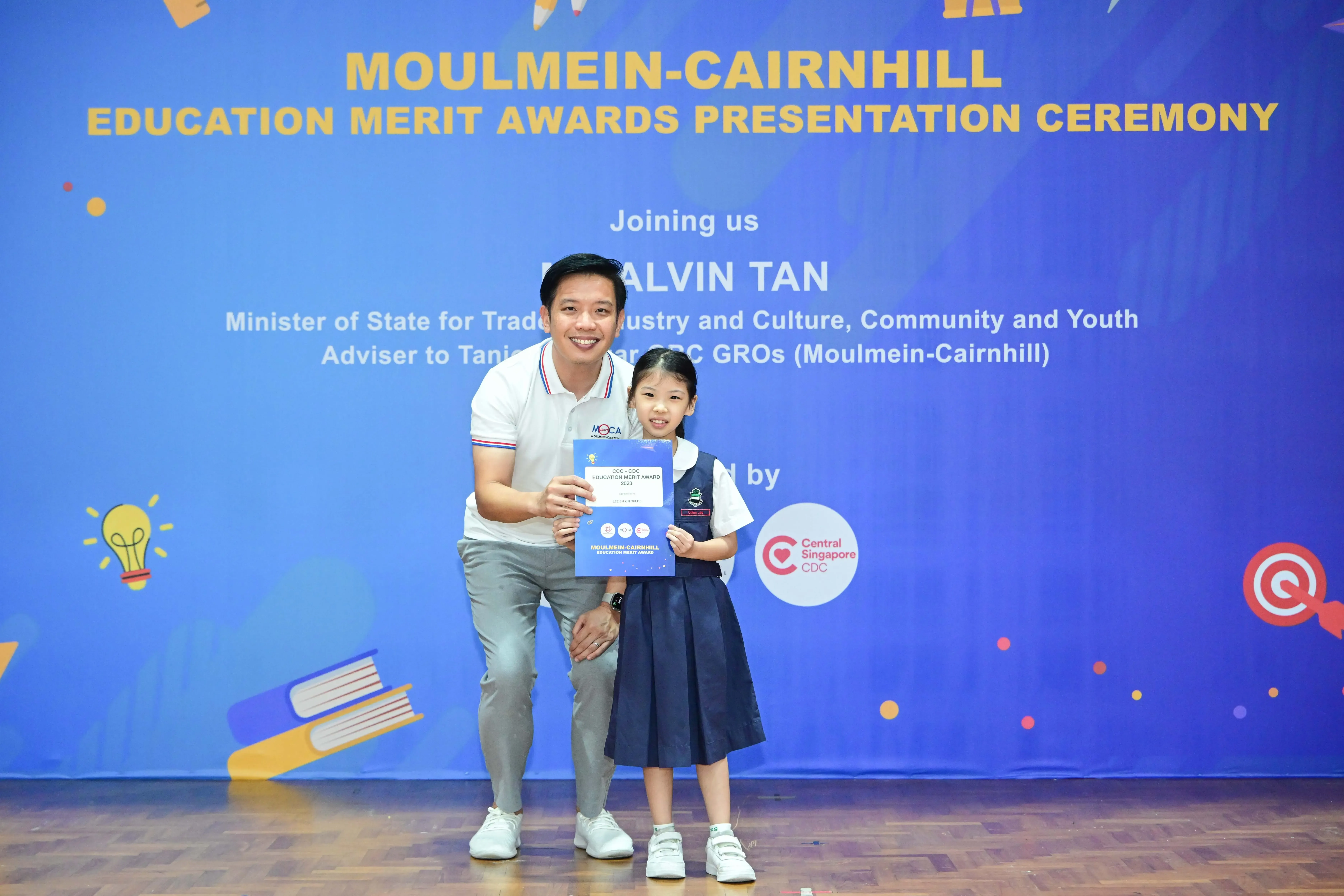 A man and a young girl in school uniform hold a certificate in front of a Moulmein-Cairnhill Education Merit Awards banner.