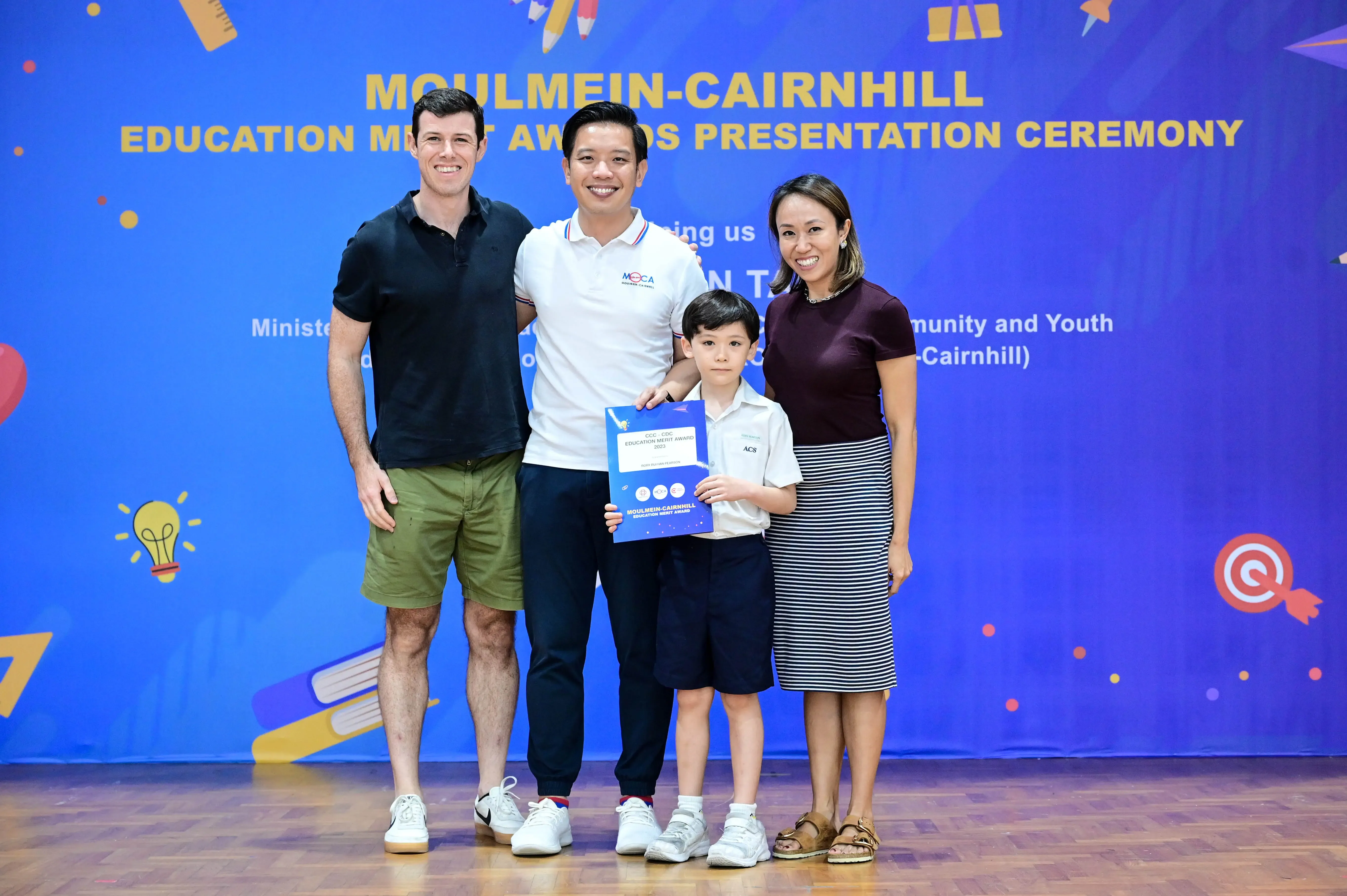 Three adults and a young boy pose at the Moulmein-Cairnhill Education Merit Awards Presentation Ceremony, boy holds award.