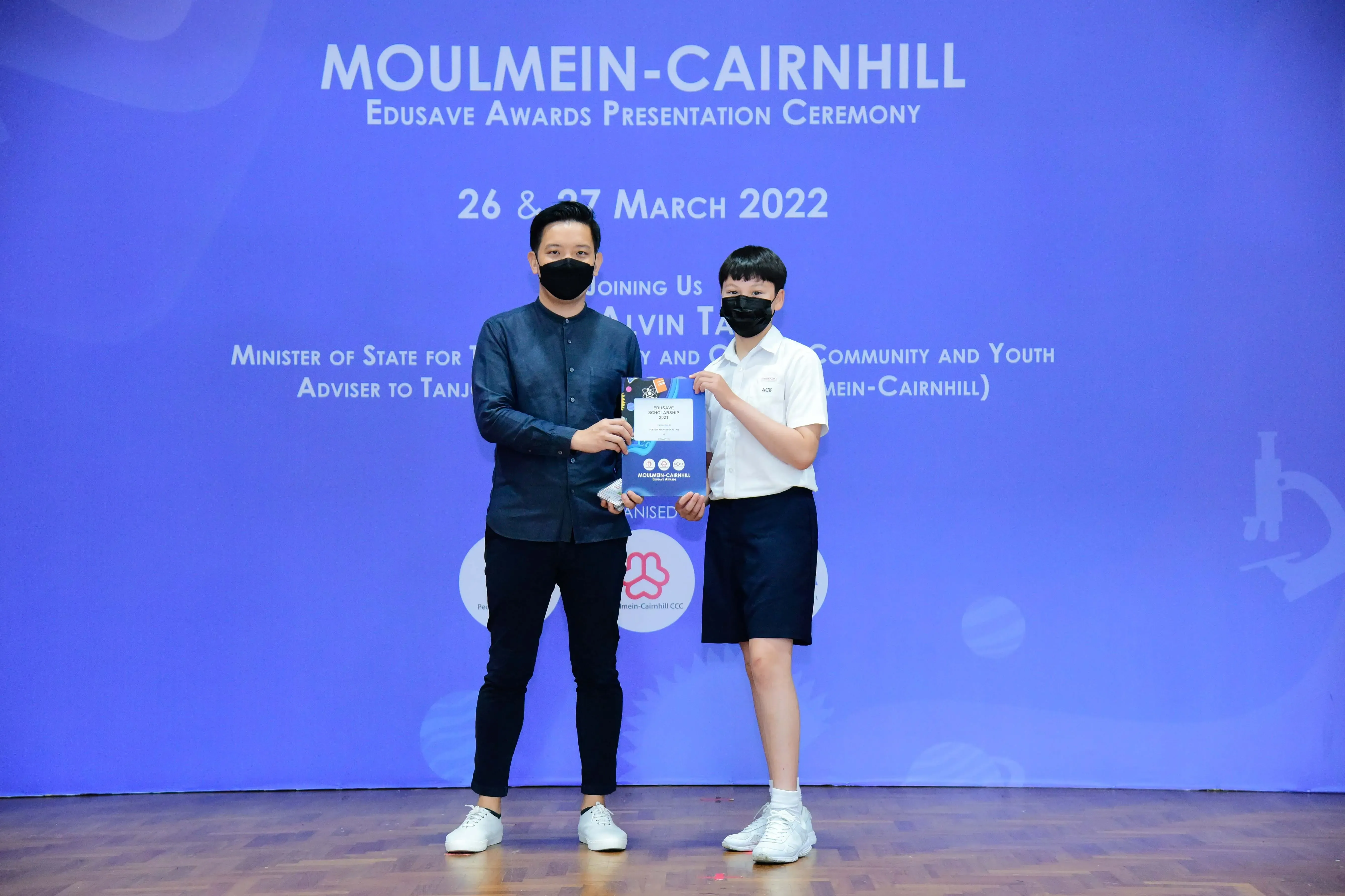 Two masked individuals hold a certificate at the Moulmein-Cairnhill Edusave Awards Presentation Ceremony, March 2022.