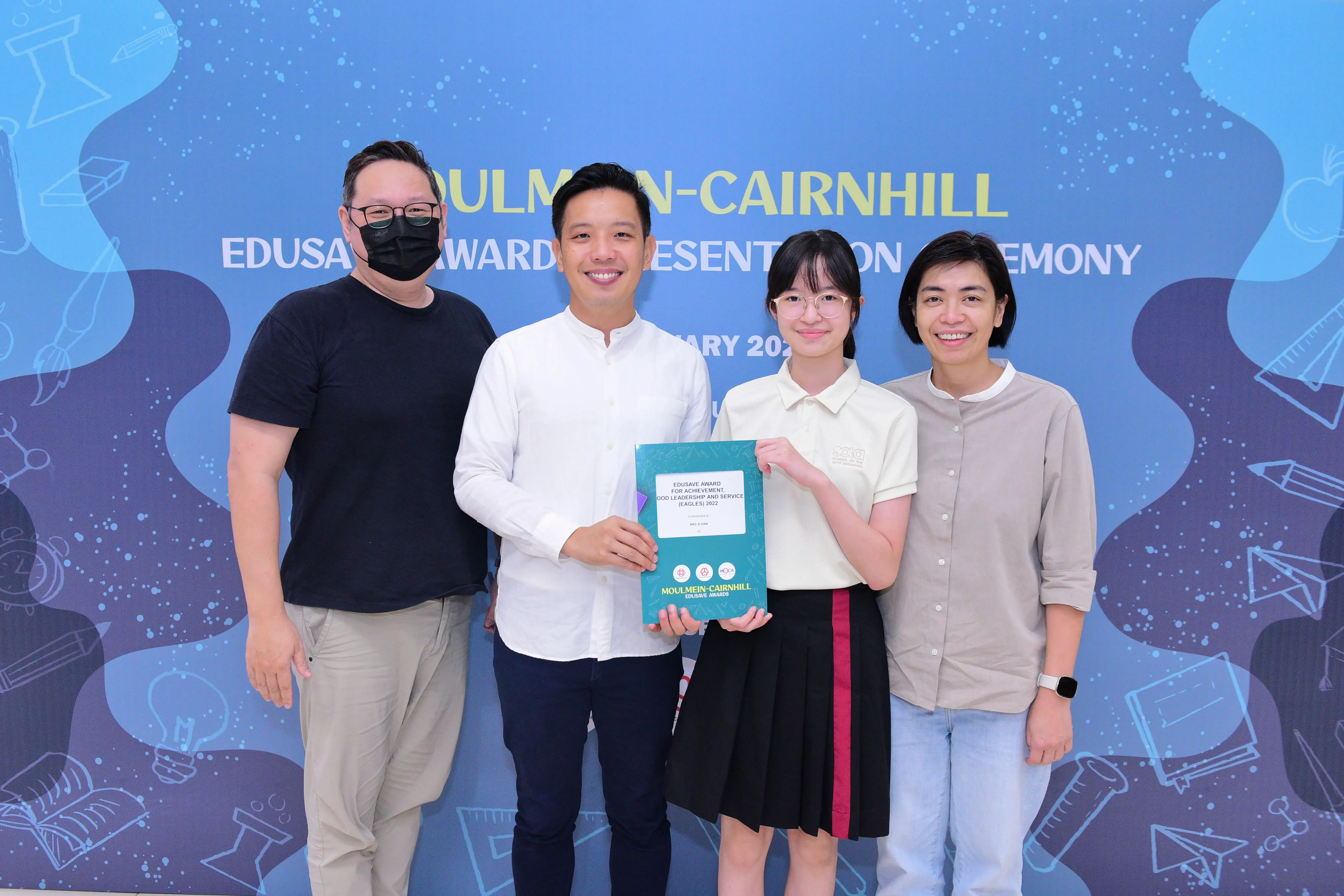 Four people pose in front of a Moulmein-Cairnhill Edusave Awards banner; a student holds a teal certificate folder.